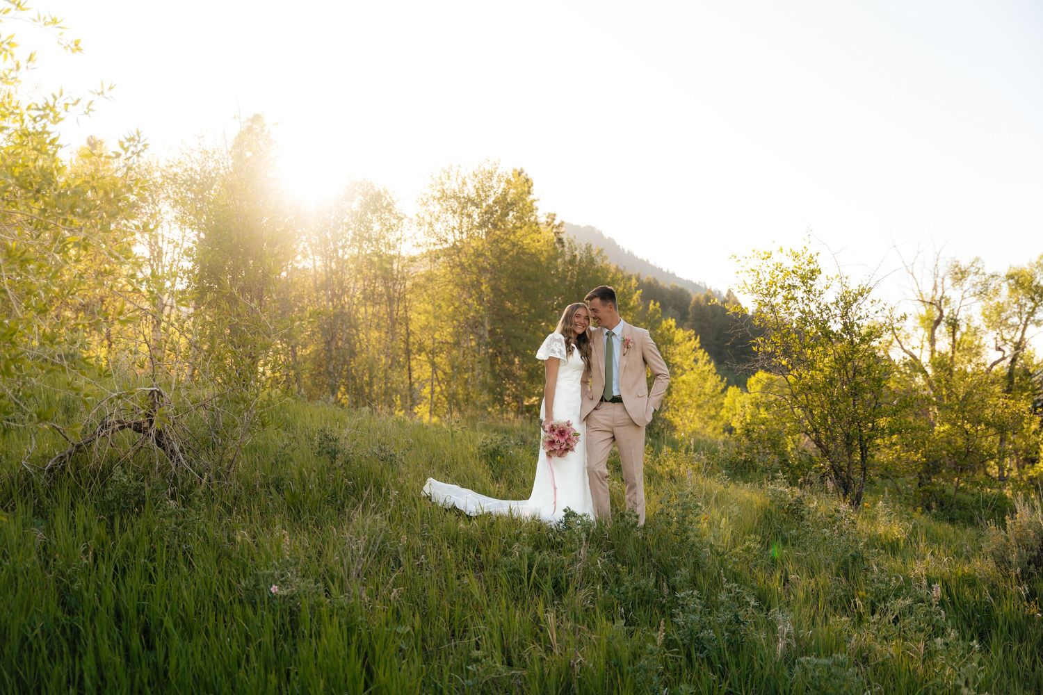 Wedding couple stands together in a green meadow with golden sunset light and mountain backdrop.