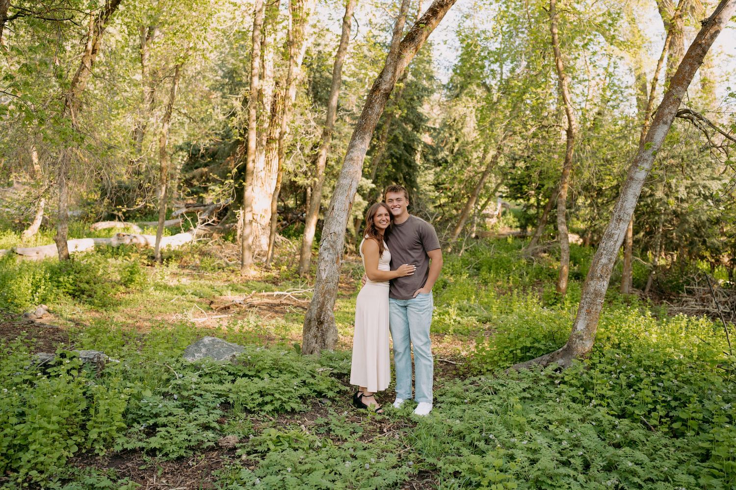 A couple embraces and smiles in a lush green forest clearing with tall trees and wildflowers.
