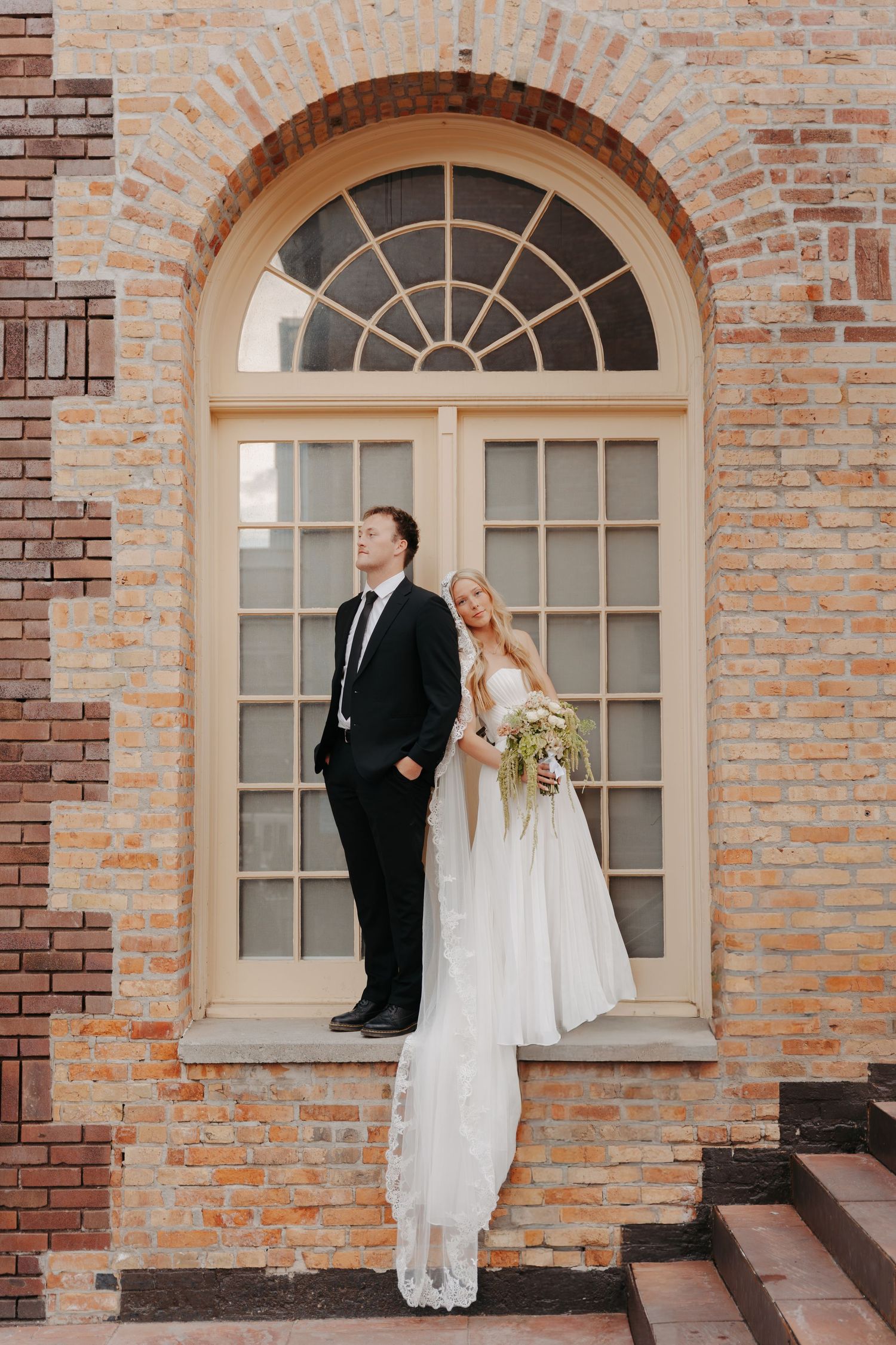 A romantic wedding couple poses against a rustic brick wall with elegant arched windows at a historic venue.