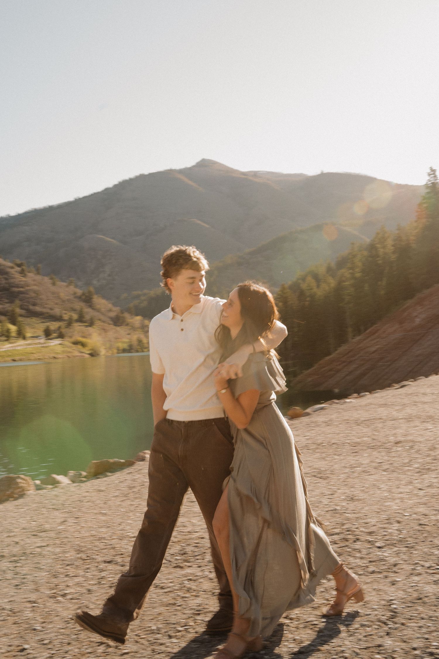 Couple walking together by a mountain lake at golden hour, woman in flowing dress embracing man in white shirt.