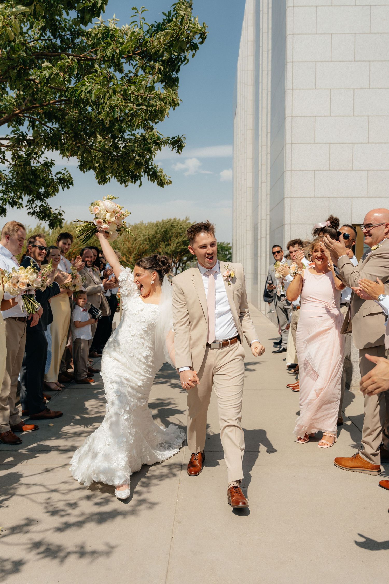 Bride and groom walk through cheering guests outdoors, bride in lace dress raising bouquet, groom in tan suit smiling.