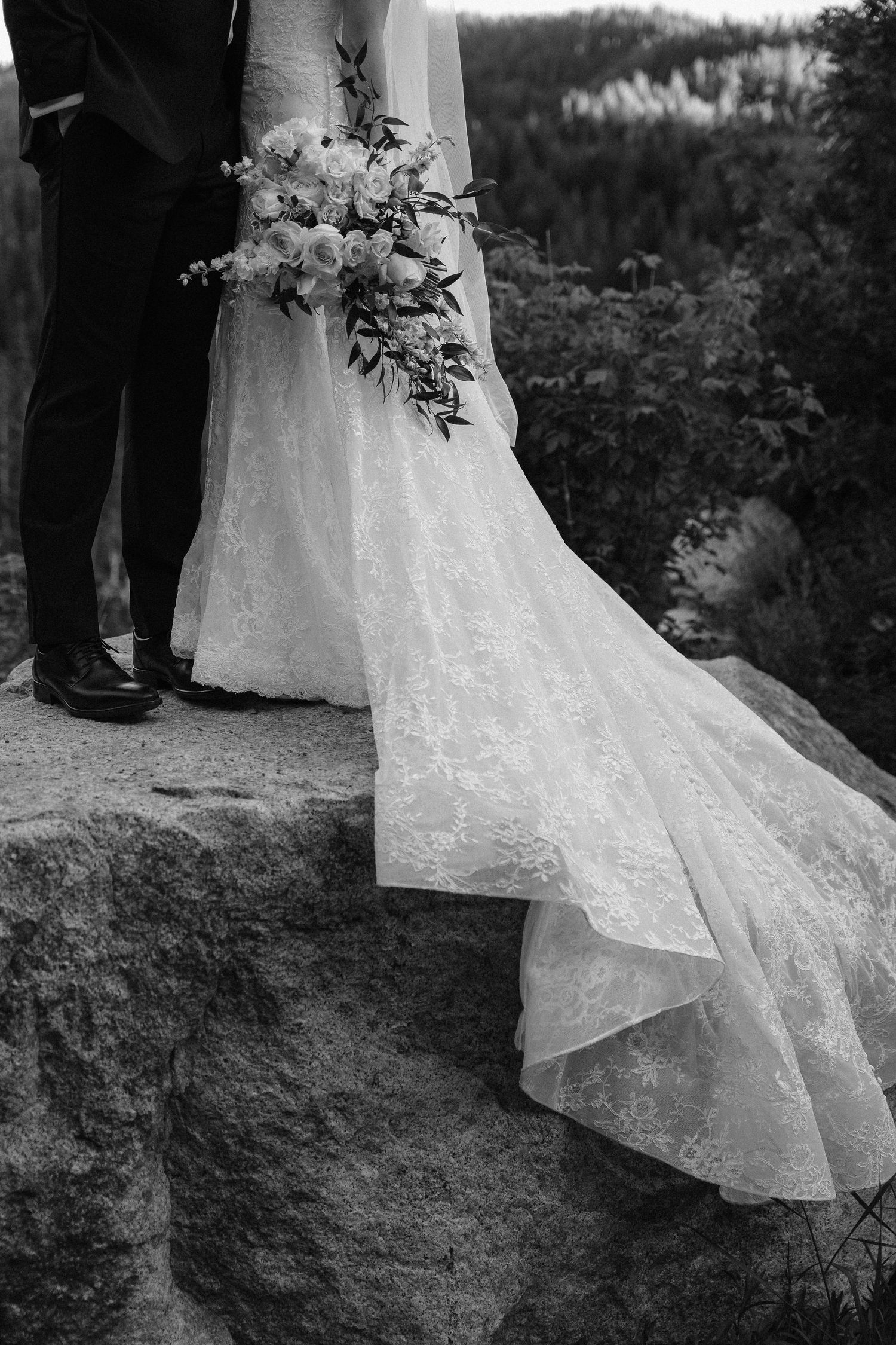 Black and white photo of bride and groom standing on rocky ledge, bride holding floral bouquet with lace dress train flowing.