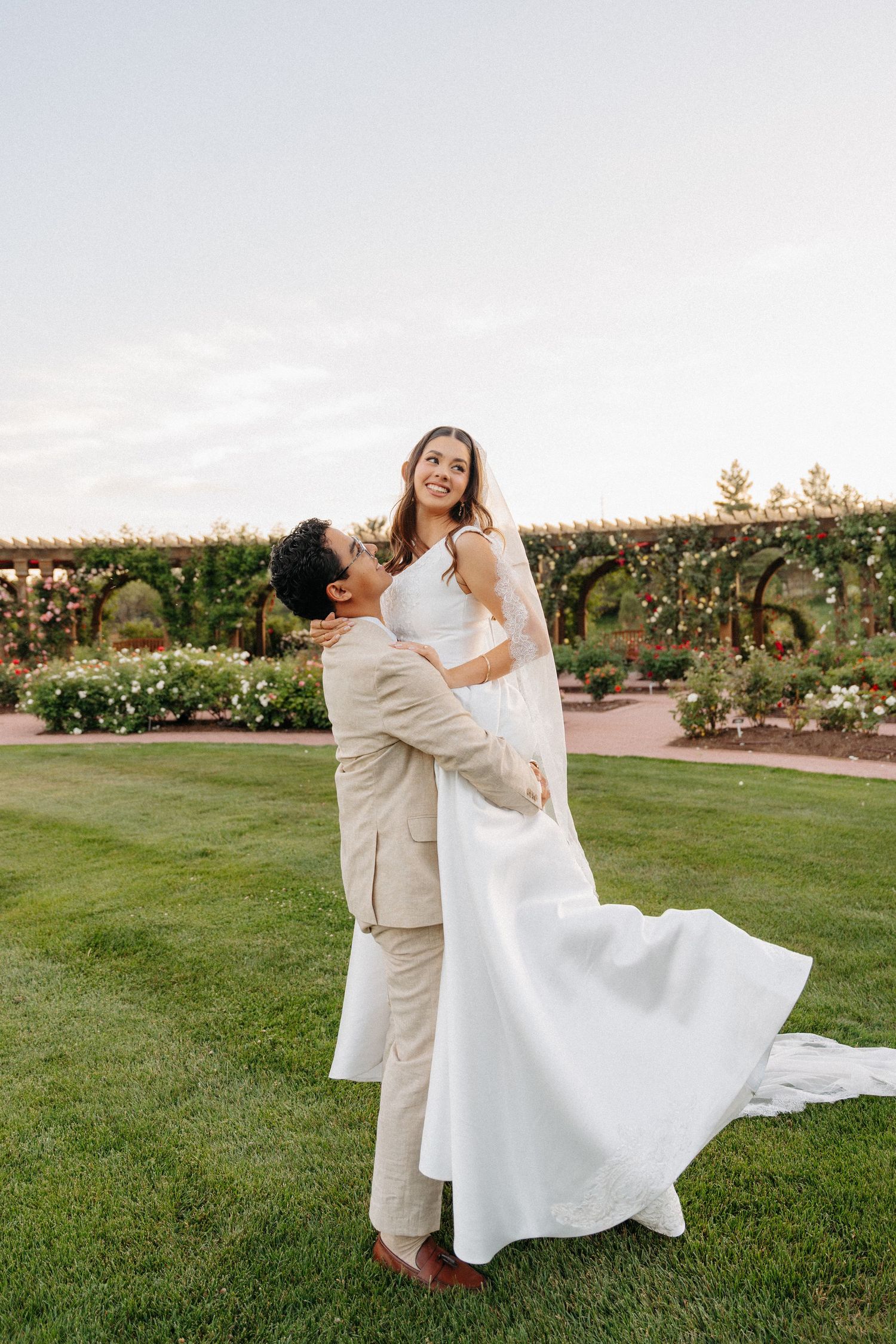 Groom lifting bride in white gown outdoors at garden wedding venue with pergola and roses in background.