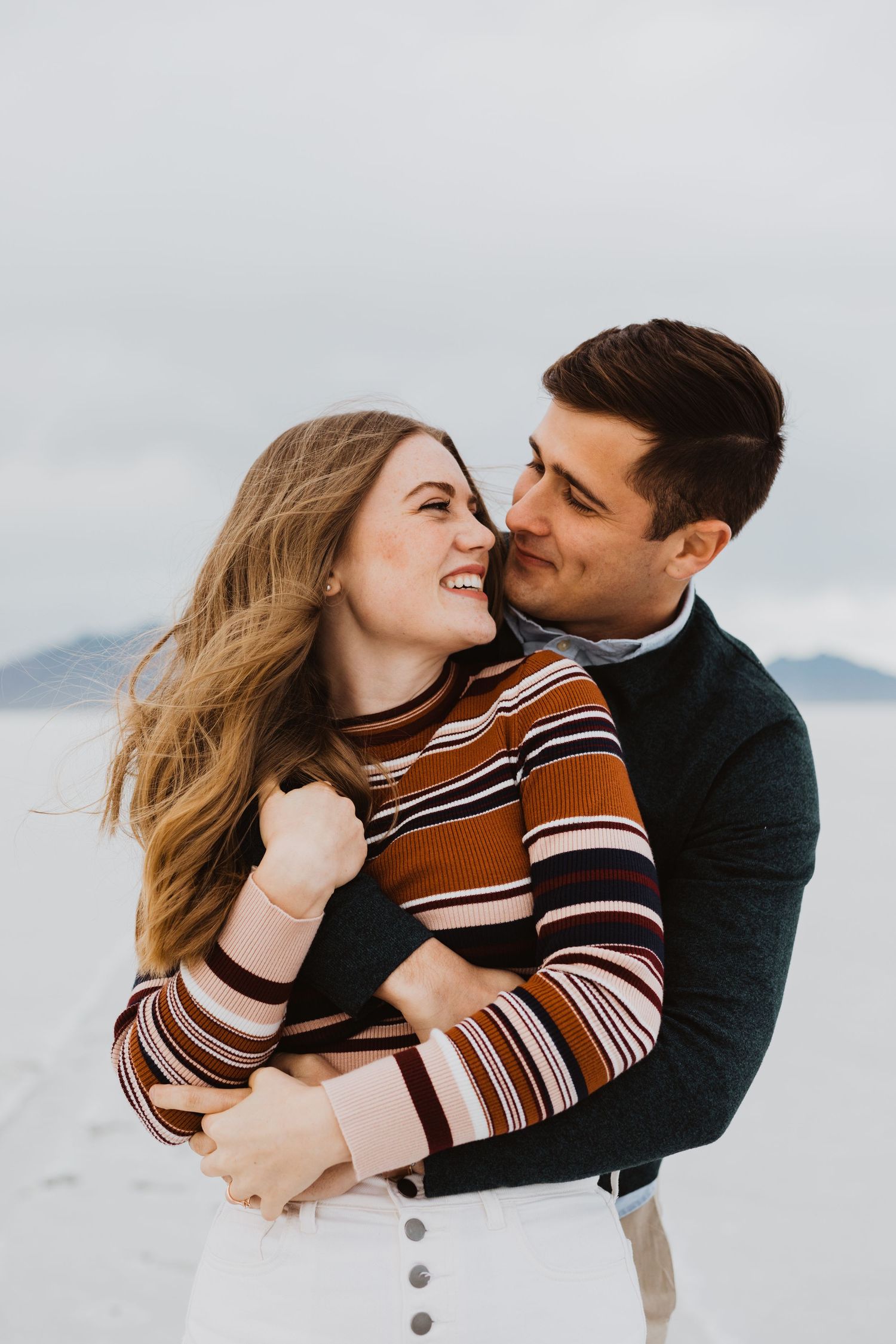 A couple embraces in striped sweater and dark jacket against a misty winter backdrop.
