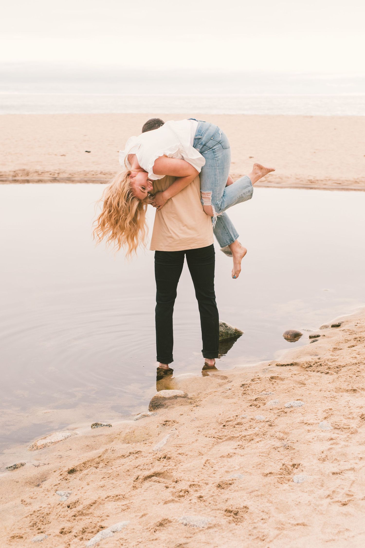 Playful couple embracing on a sandy beach at sunset with soft pastel tones.