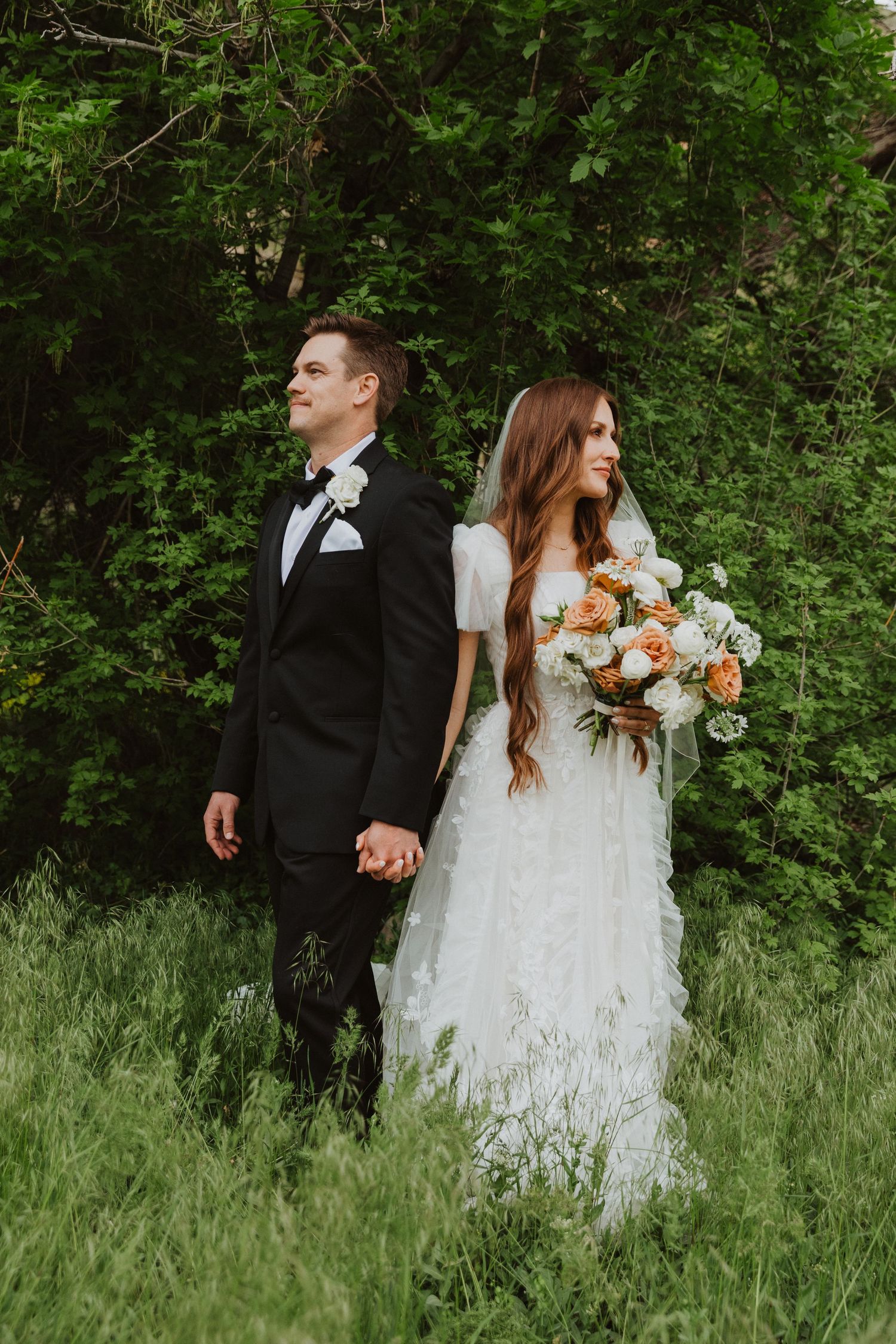 A couple in formal wedding attire poses in a lush green forest setting.