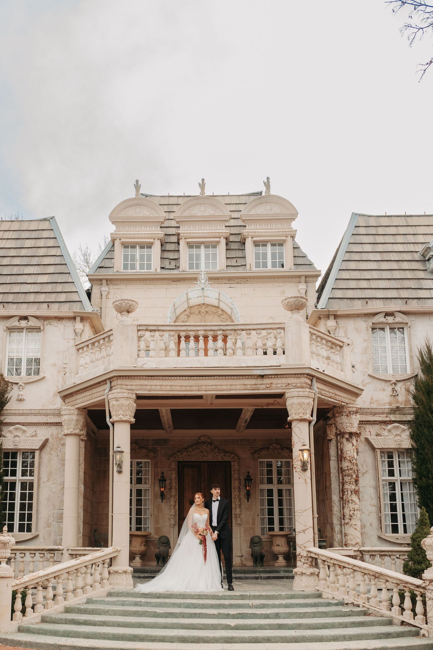 Elegant wedding couple poses in front of a grand French-style mansion with ornate architecture.