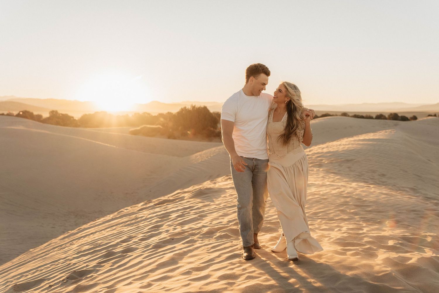 A couple walks hand in hand through sandy dunes at sunset creating long shadows across the desert landscape.