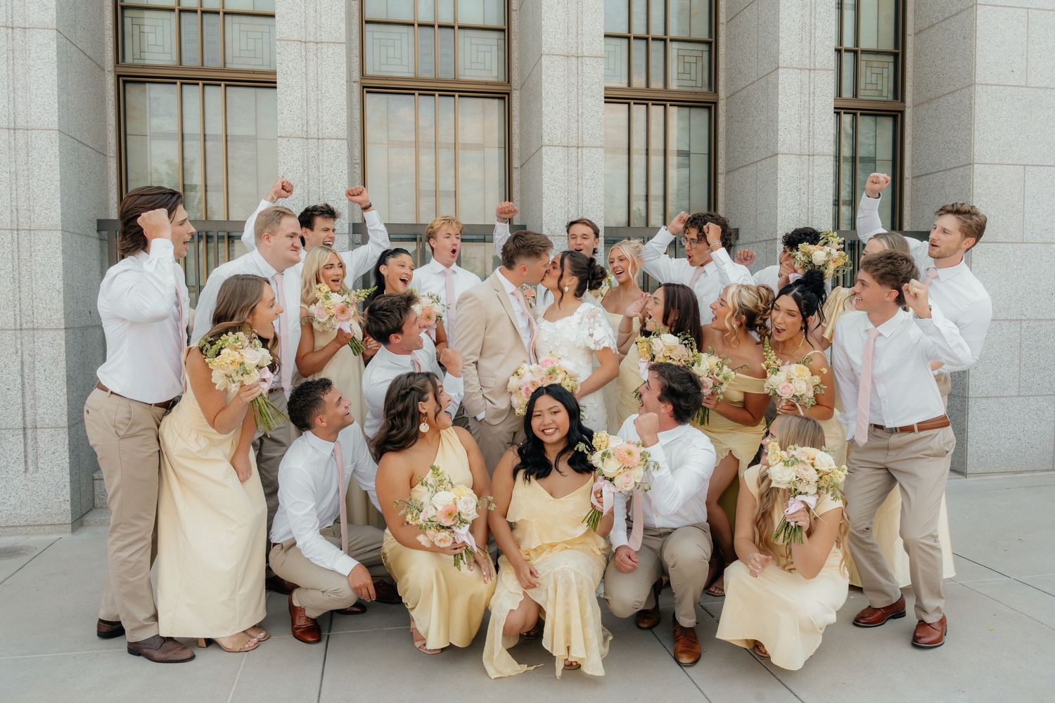 Large wedding party celebrating outdoors in cream and yellow attire, holding bouquets and cheering on venue steps.