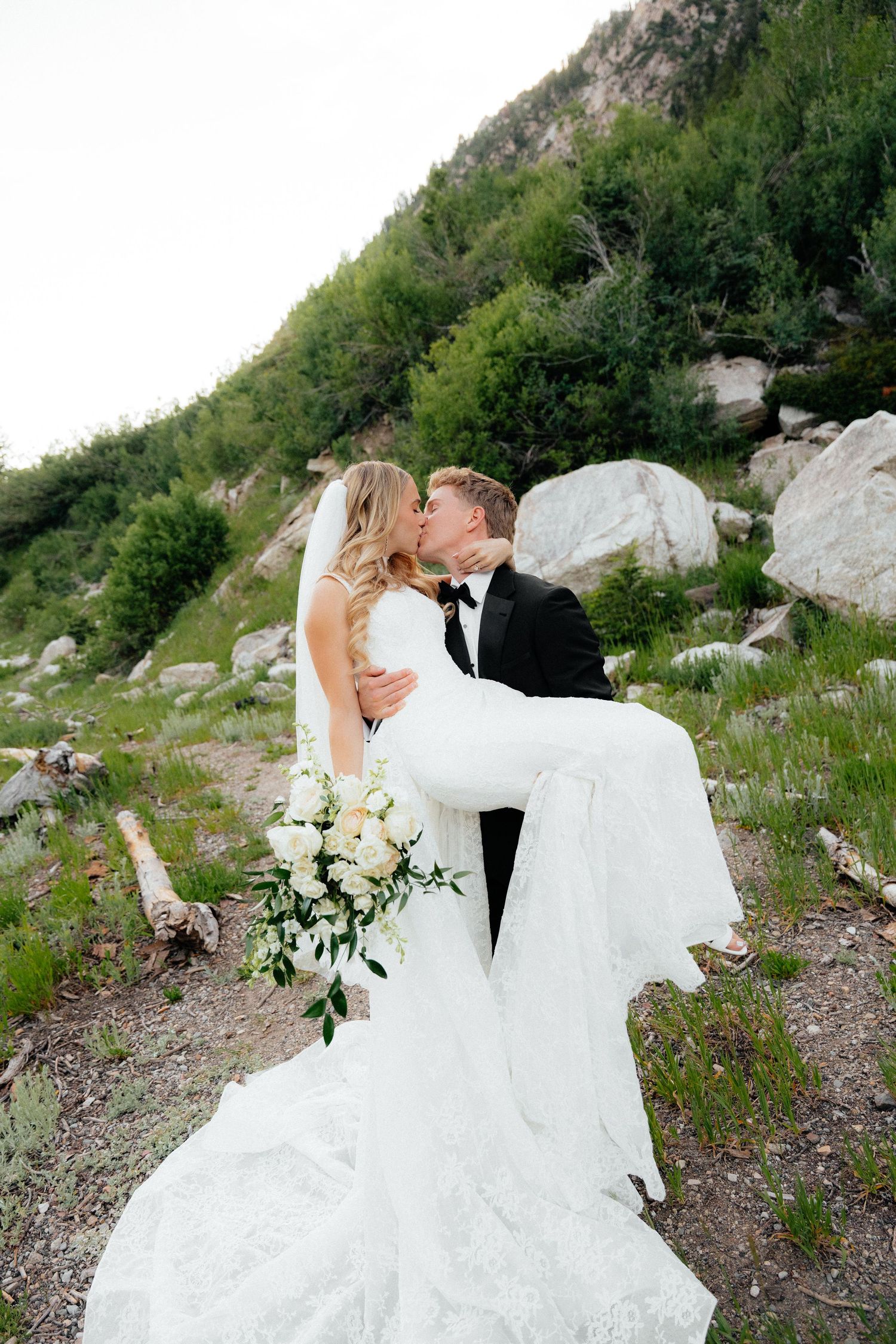 Groom in black tuxedo lifts bride in white lace gown as they kiss on a rocky mountain trail with greenery.