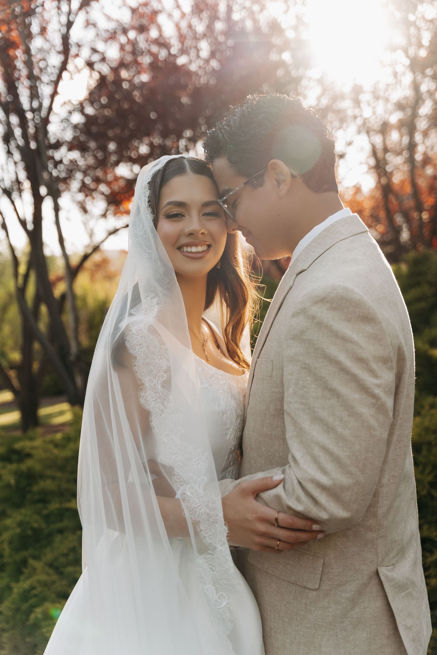 Bride in lace veil smiles as groom in beige suit embraces her during golden hour outdoor wedding portrait.