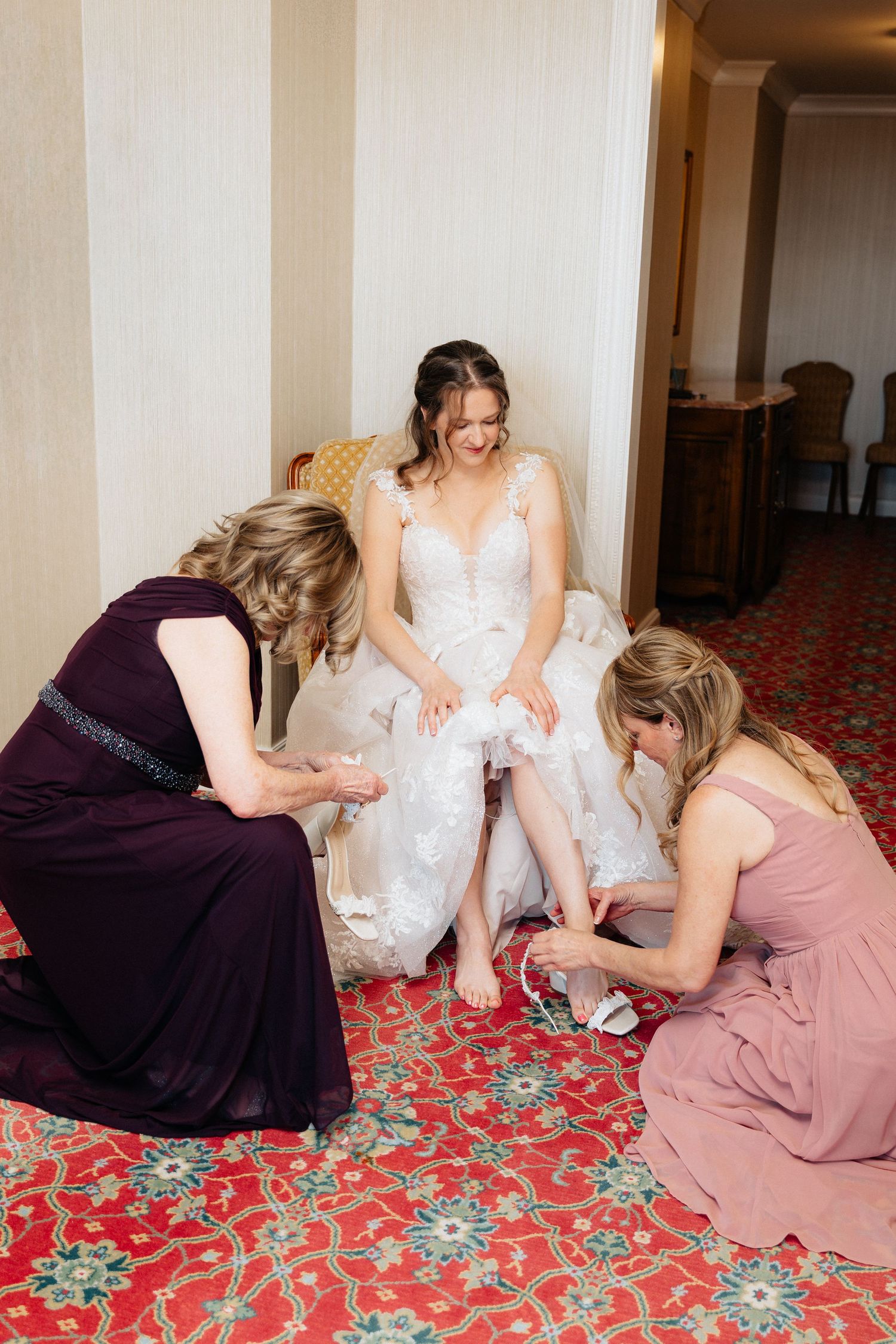 Bride in white gown being helped into shoes by two bridesmaids in purple and pink dresses in elegant hotel room.