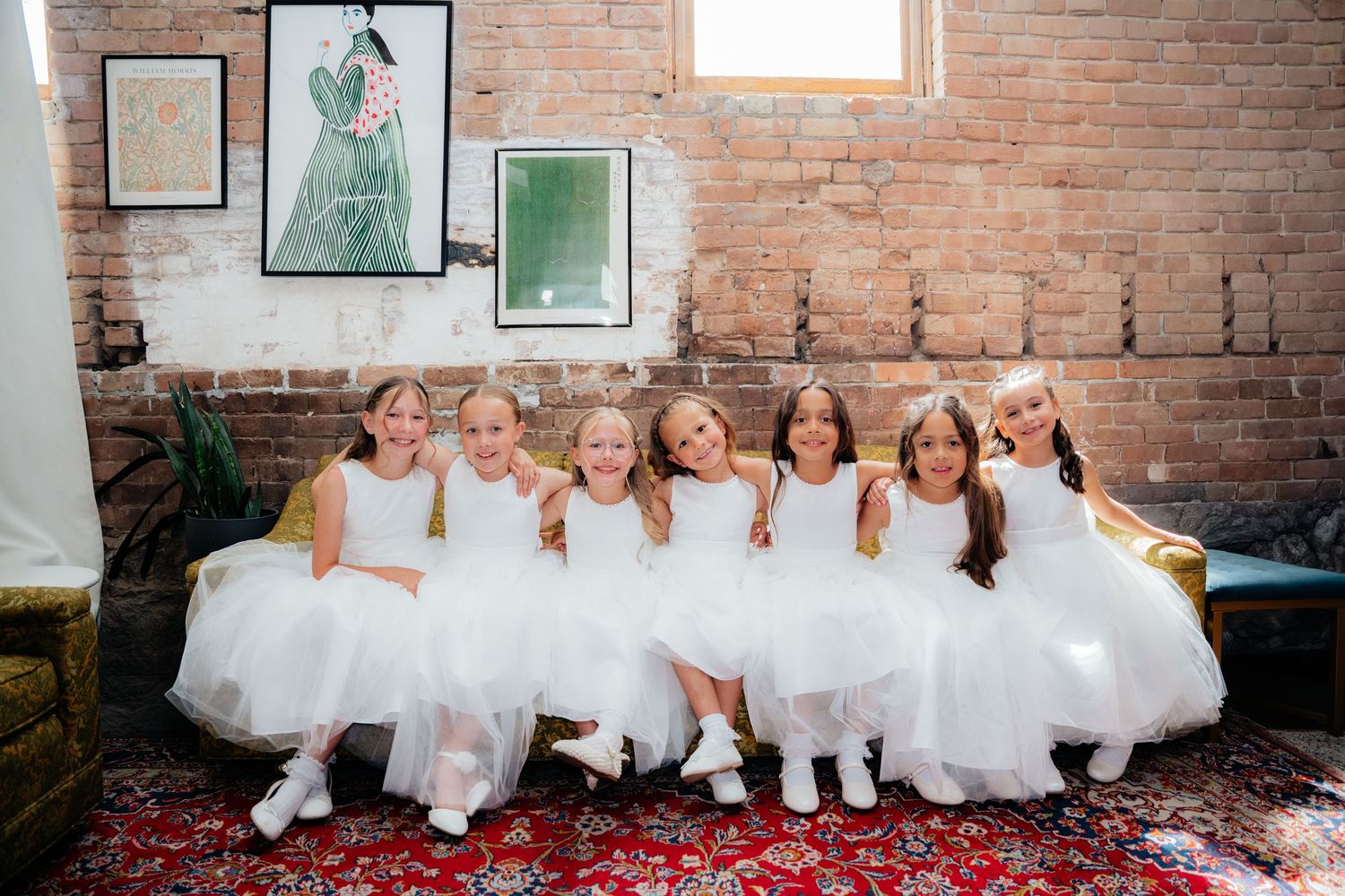 Seven flower girls in white dresses sitting together on a vintage sofa, smiling at a wedding venue with brick walls.
