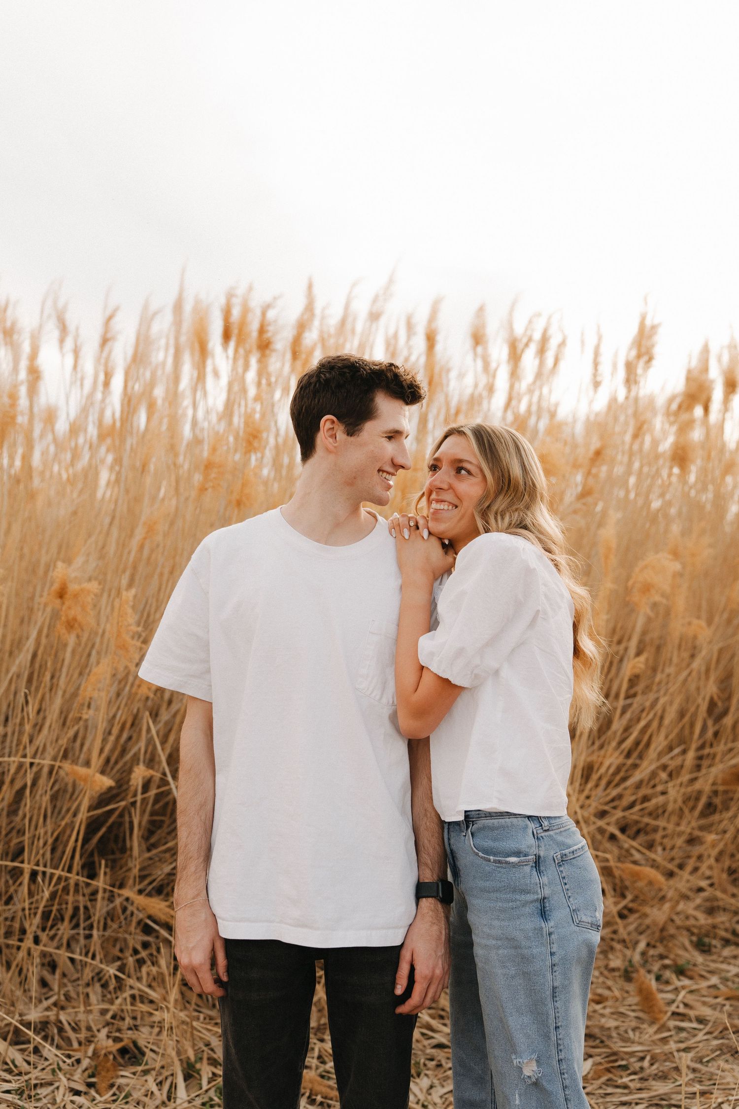A young couple in matching white tops stands smiling in a golden wheat field, the woman resting her hand on the man's chest.