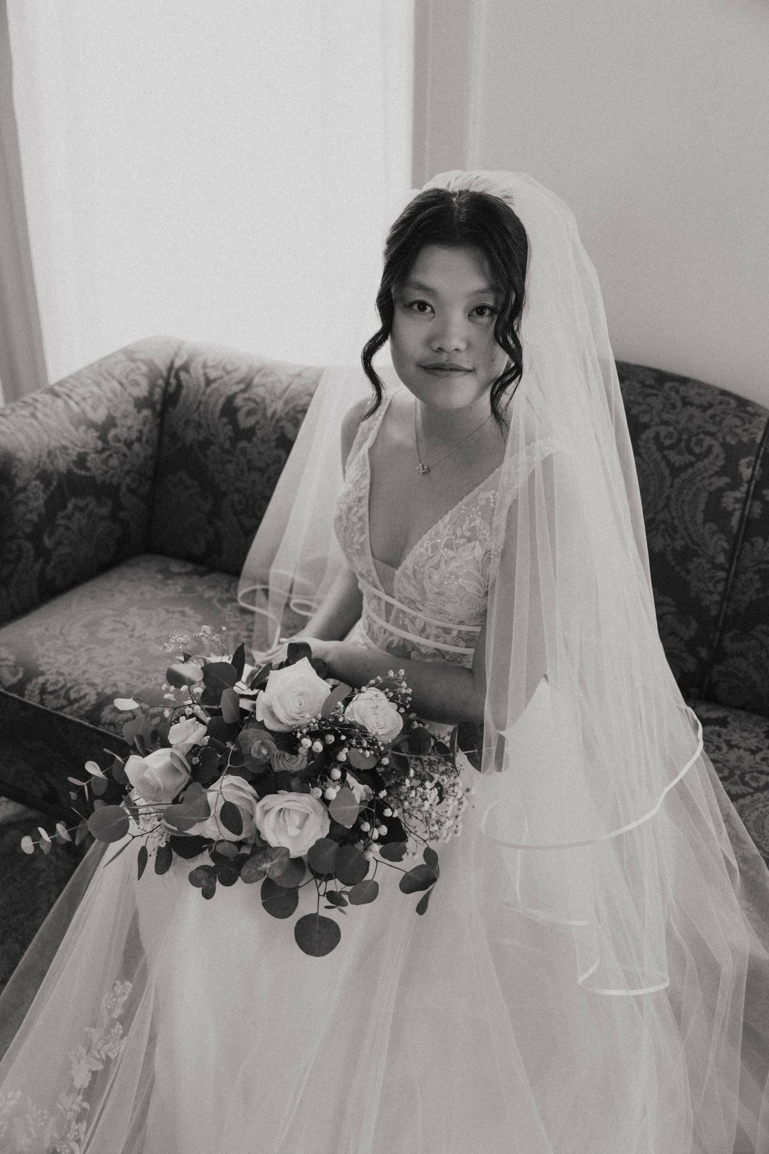 Black and white photo of a bride in a lace gown and veil holding a bouquet while seated on a floral sofa.