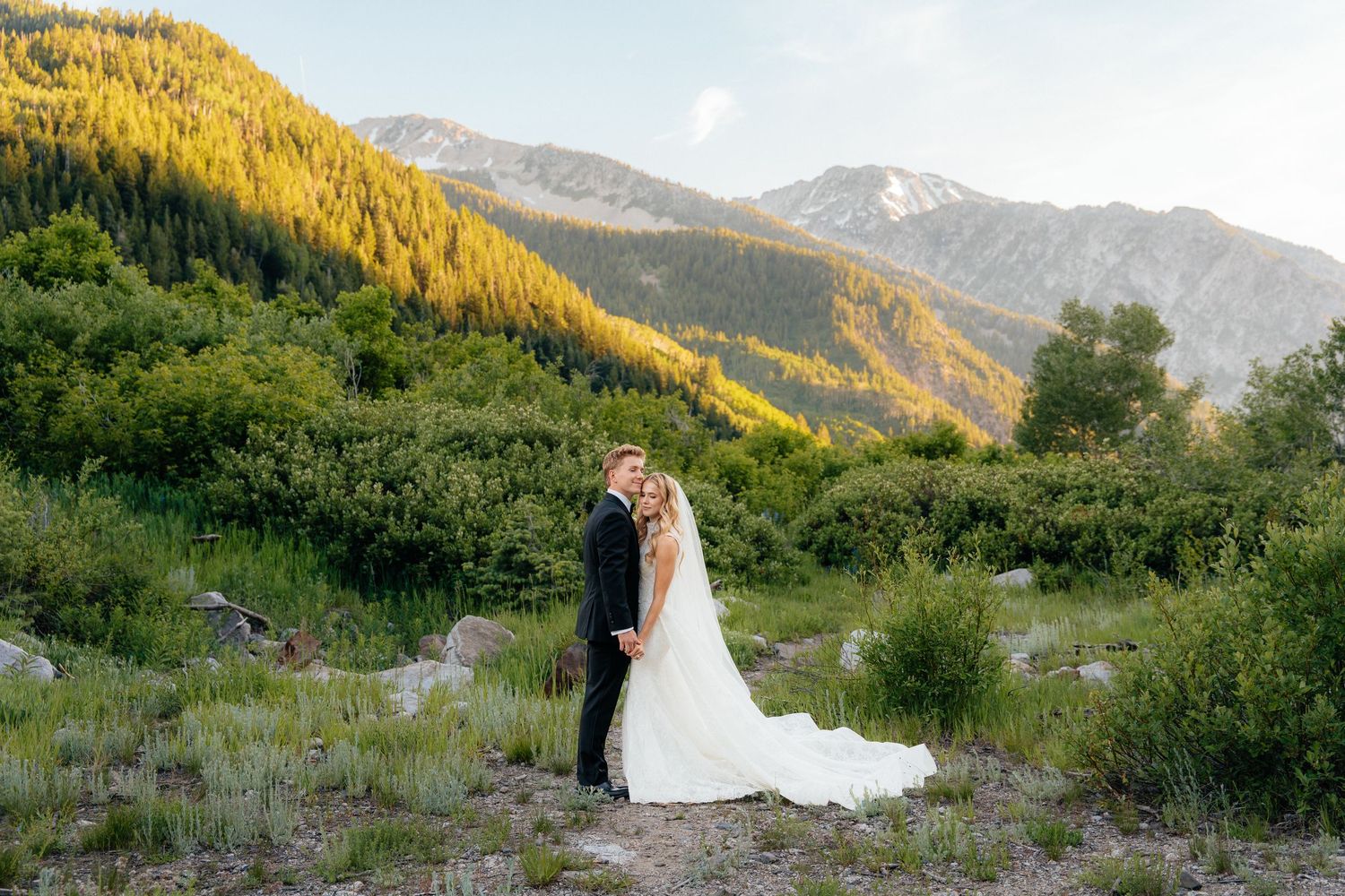 Bride and groom embrace in mountain meadow with golden sunlit peaks and lush green trees at sunset wedding portrait.