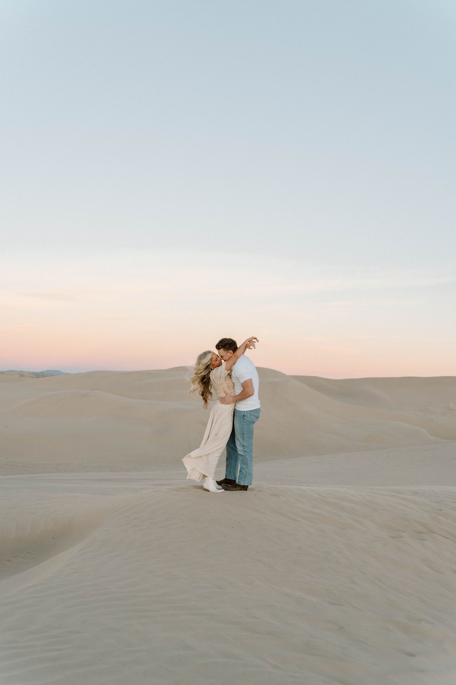 A romantic couple dances together on rolling sand dunes during sunset in a desert landscape.
