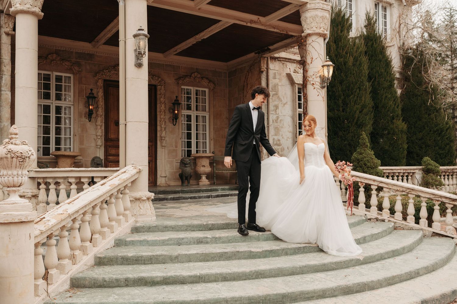 Elegant wedding couple descends grand stone staircase at historic mansion venue.