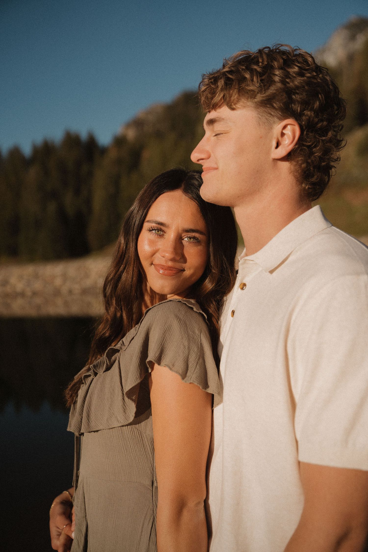 A young couple poses outdoors near a mountain lake, the woman smiling warmly as golden hour light illuminates them.