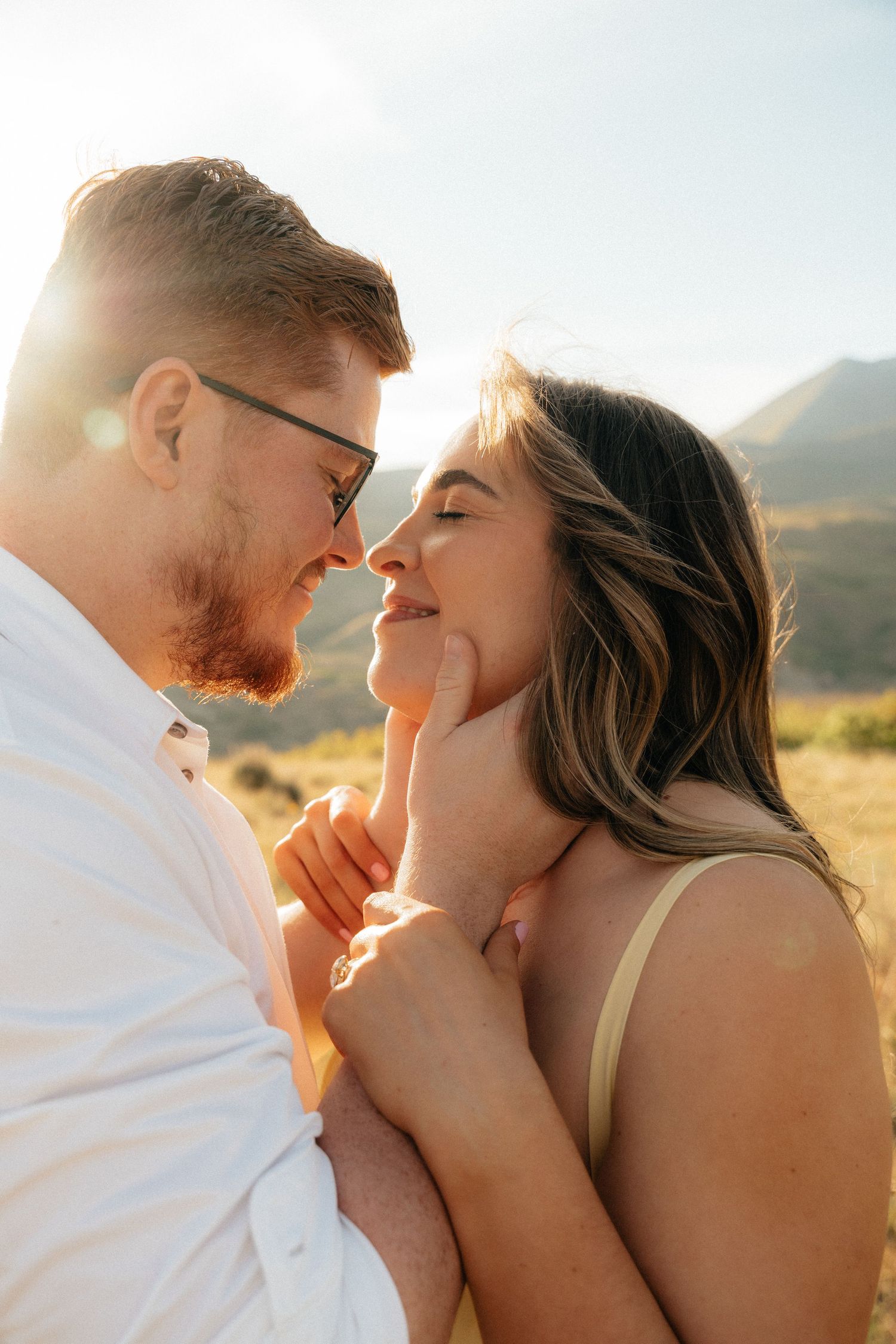A couple shares a romantic moment outdoors with golden sunlight and mountains in the background.