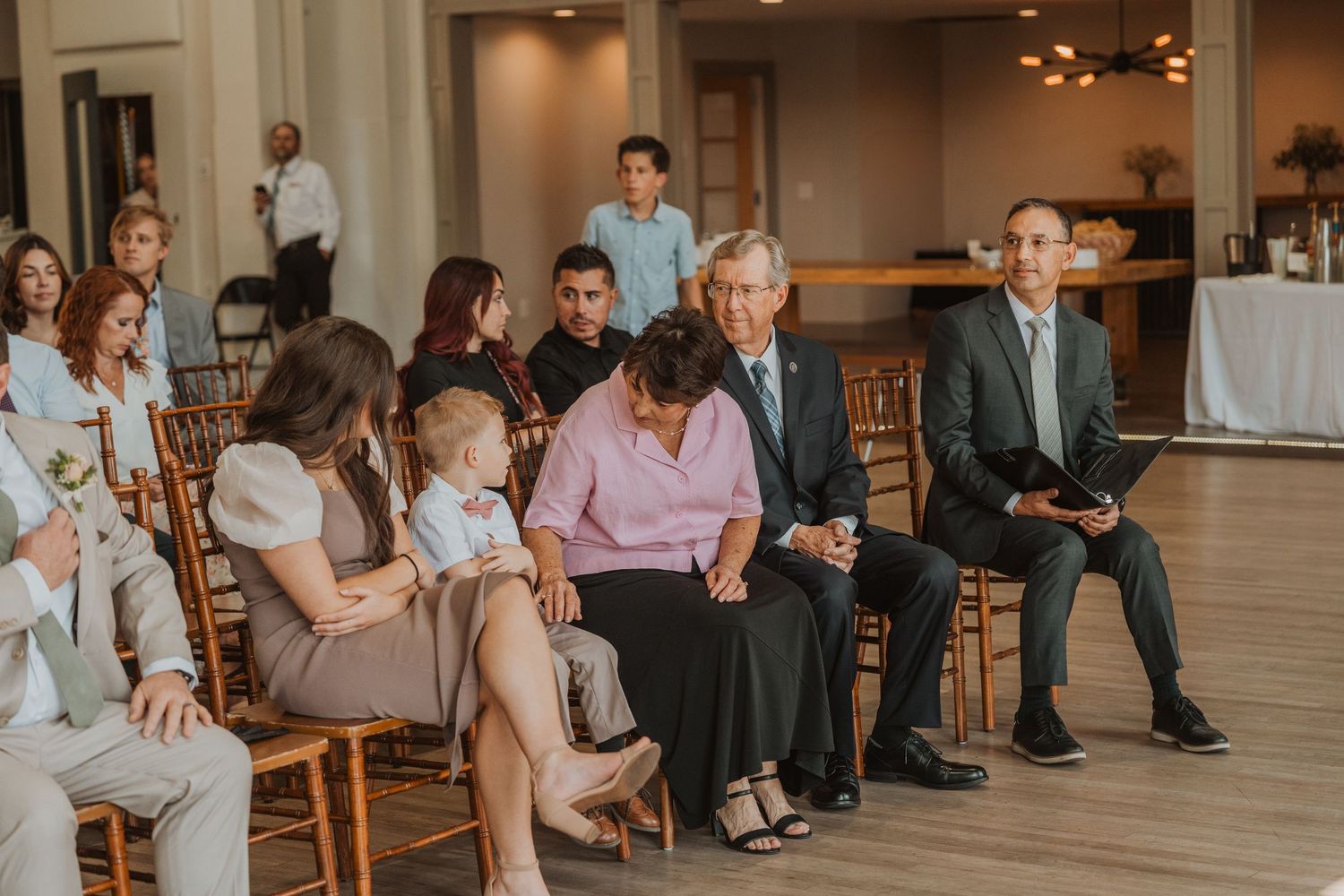 Indoor wedding ceremony seating with wooden chairs and guests in formal attire.