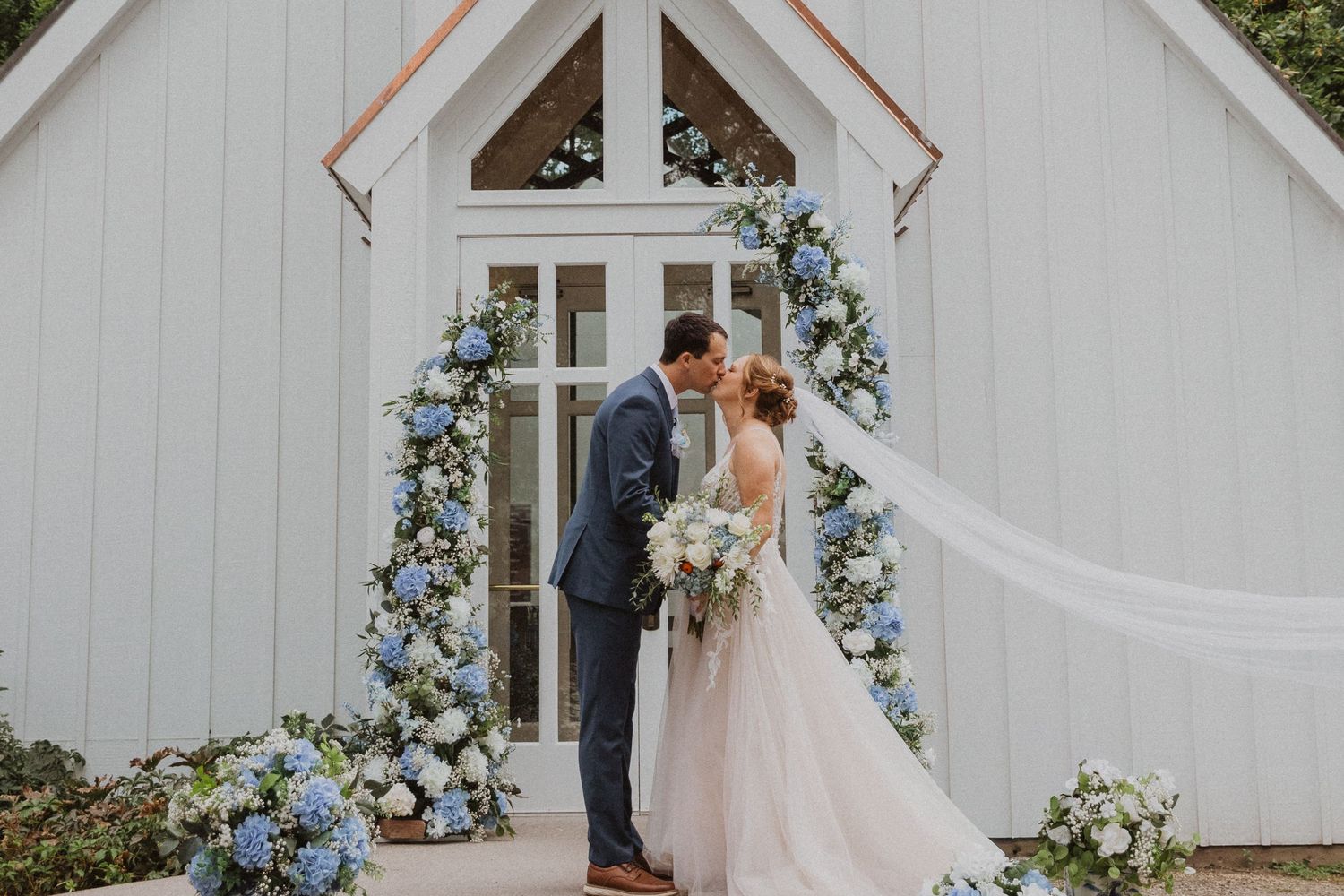 A romantic wedding ceremony Utah takes place in front of a white chapel adorned with blue and white floral decorations.