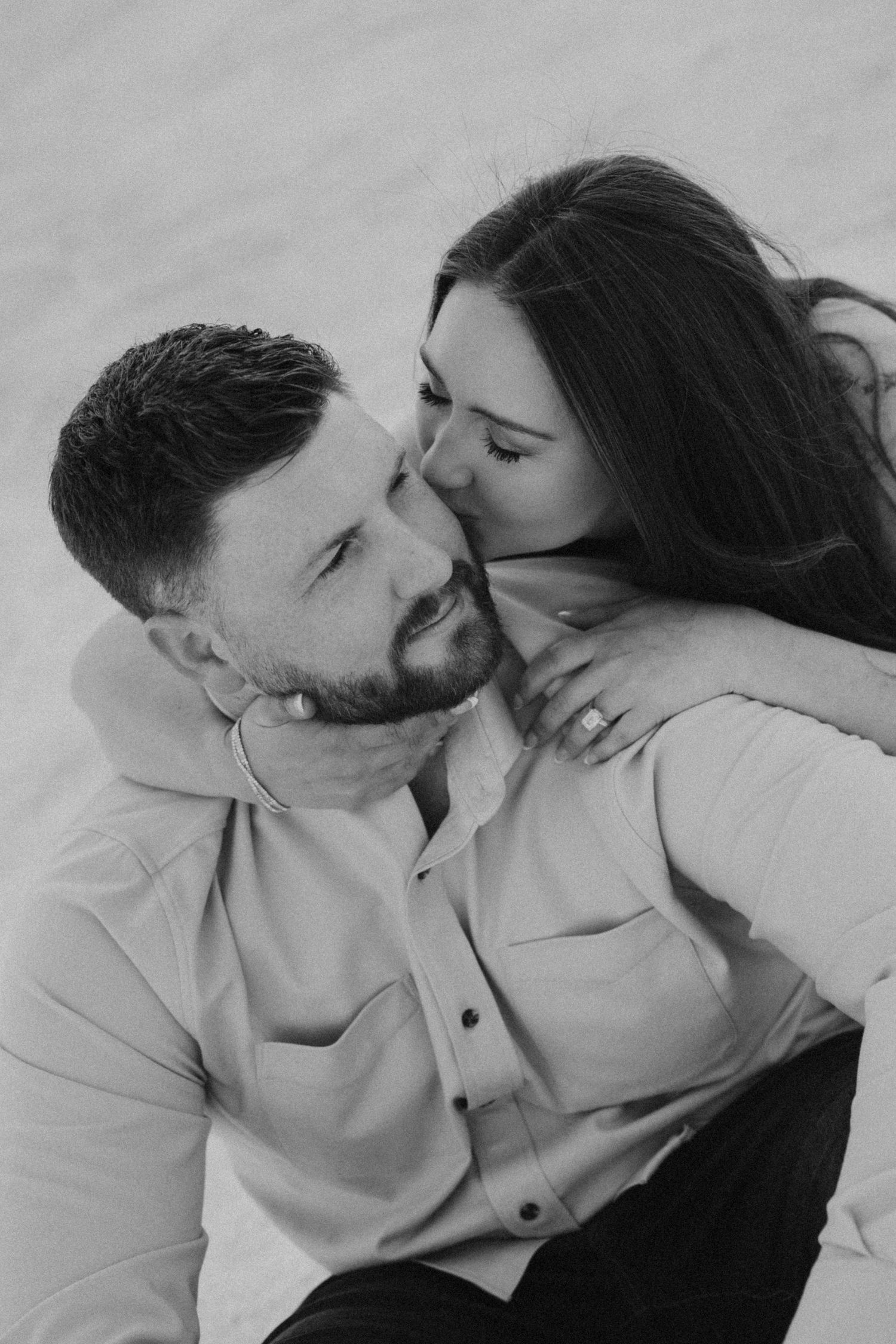 Black and white photo of woman kissing mans cheek while embracing outdoors on sandy terrain.