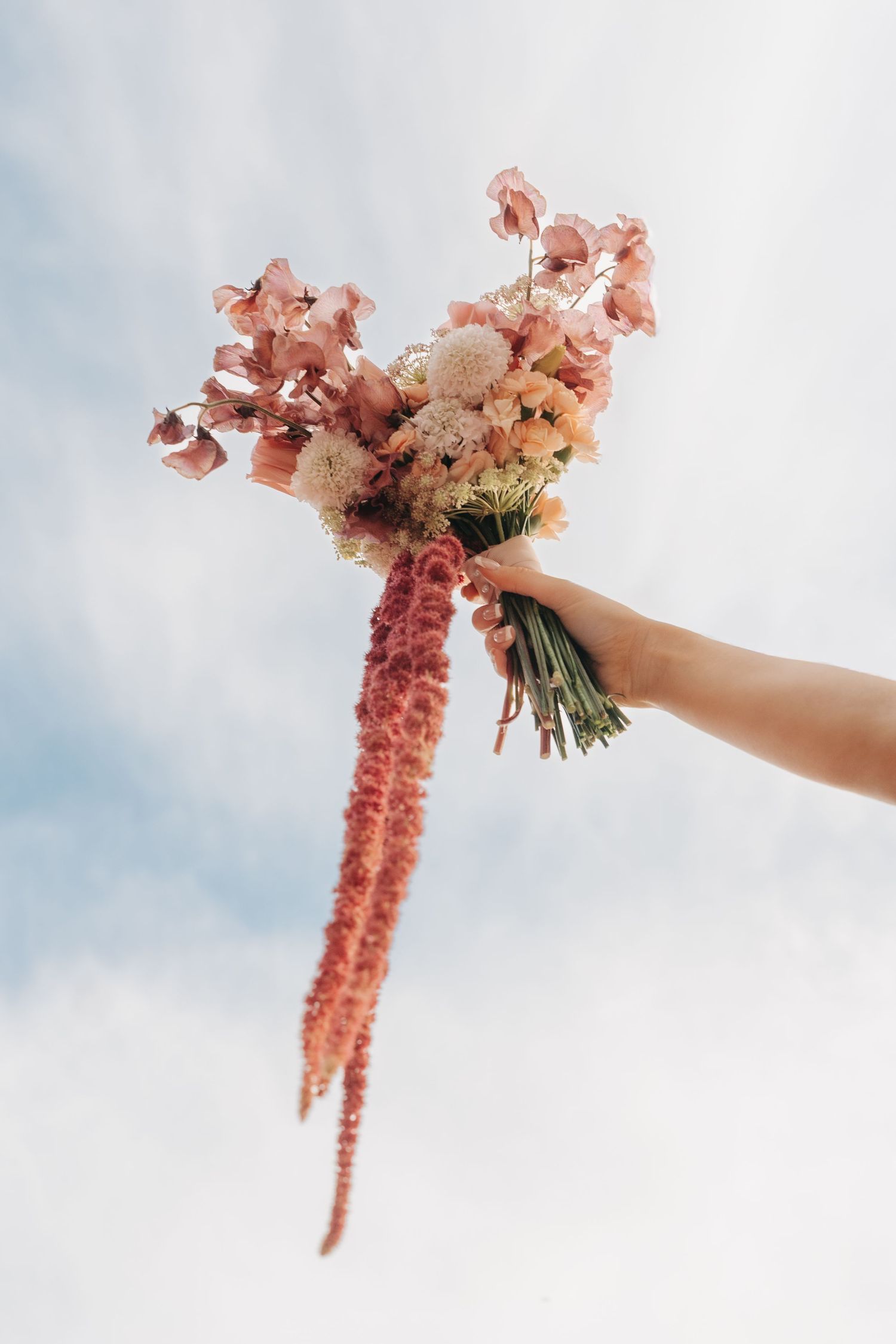 Pink and coral wedding bouquet with trailing amaranthus held against cloudy sky.