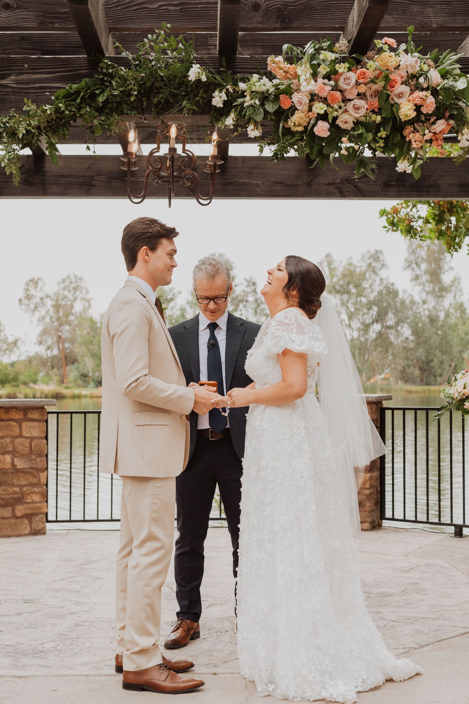 Wedding party gathers on stone patio under wooden beams decorated with peach and white flowers.