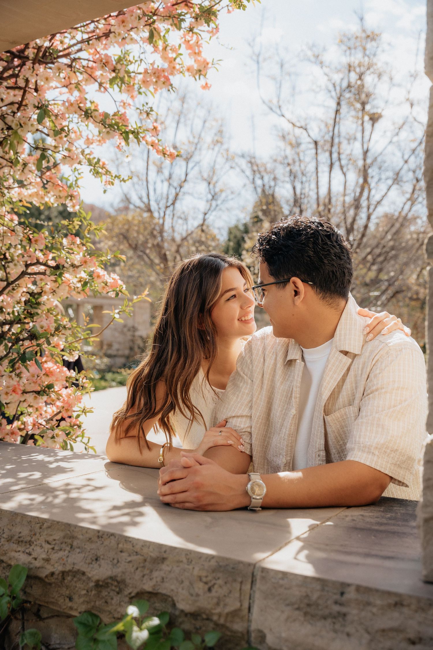 Romantic couple leaning on stone balcony surrounded by pink spring blossoms, sharing a tender moment outdoors.