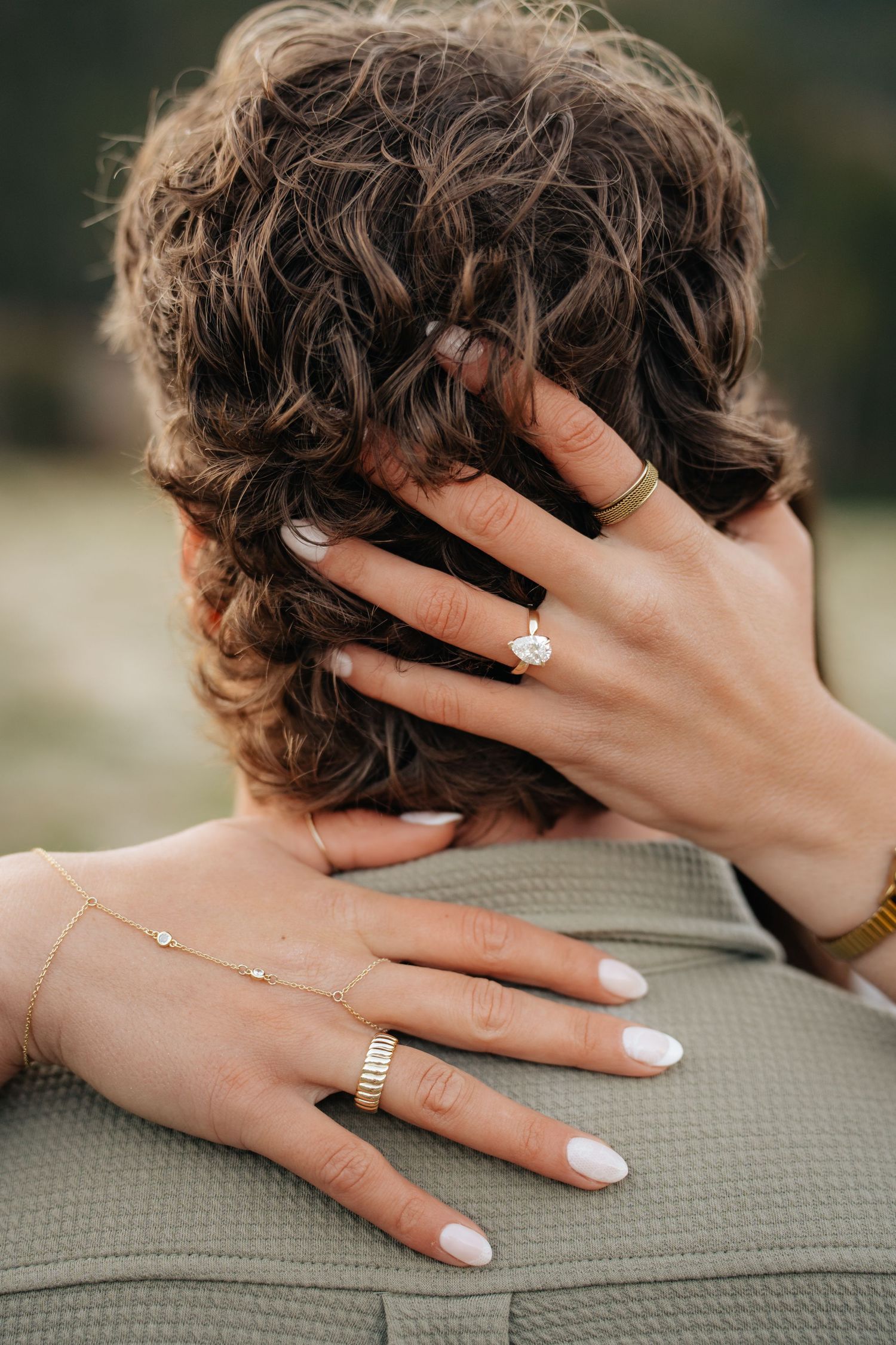 Close-up of woman's hands with engagement ring and gold jewelry resting on man's shoulders and curly hair.