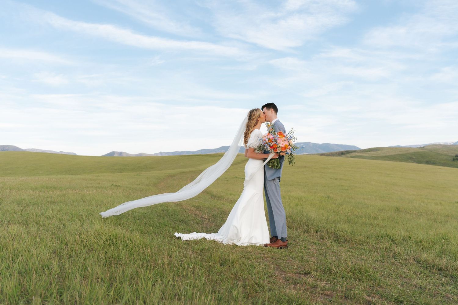 Bride and groom kissing in a lush green meadow, her veil flowing in the wind, with rolling hills in the background.