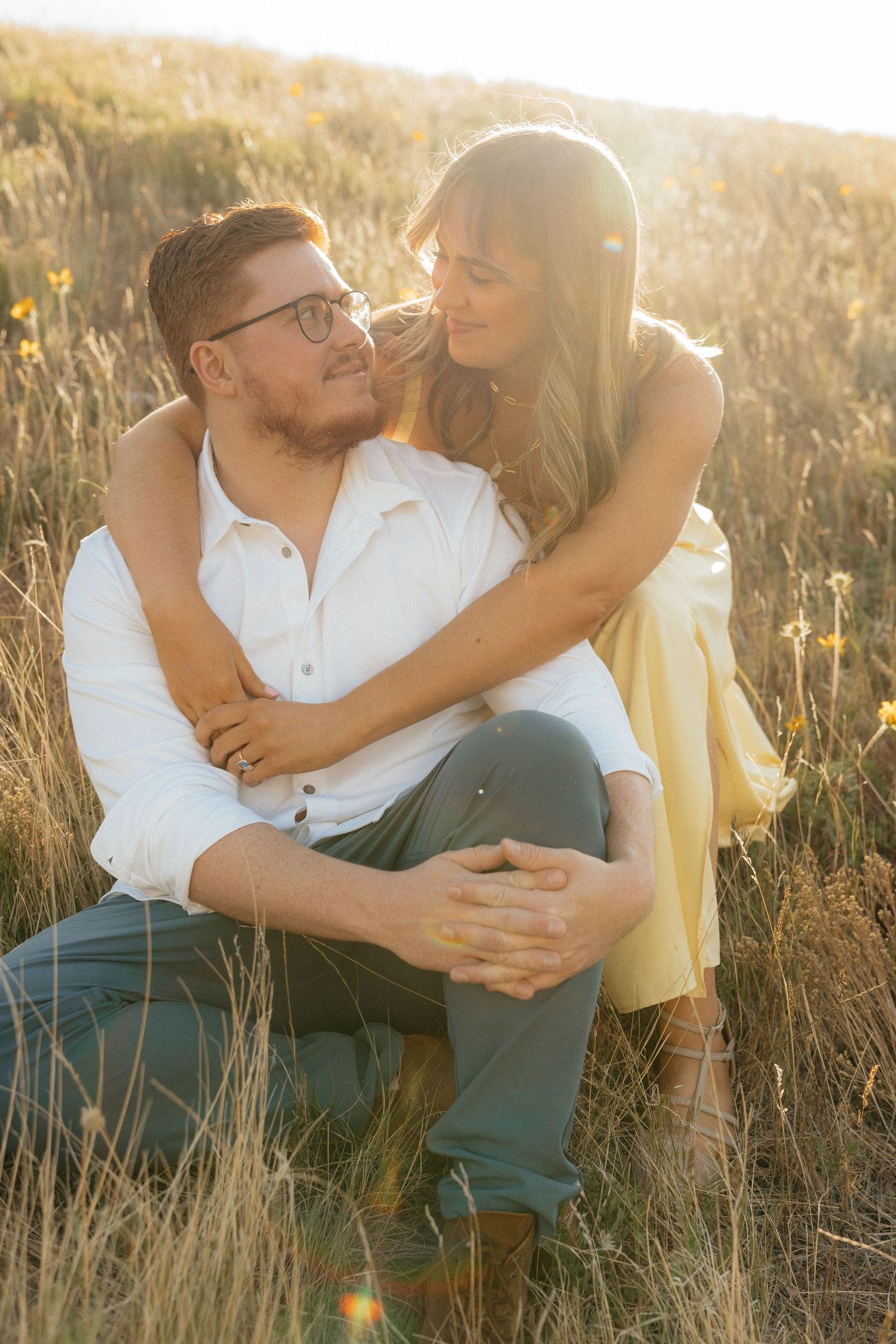 Couple embracing in golden sunlit field, woman in yellow dress hugging man in white shirt wearing glasses.