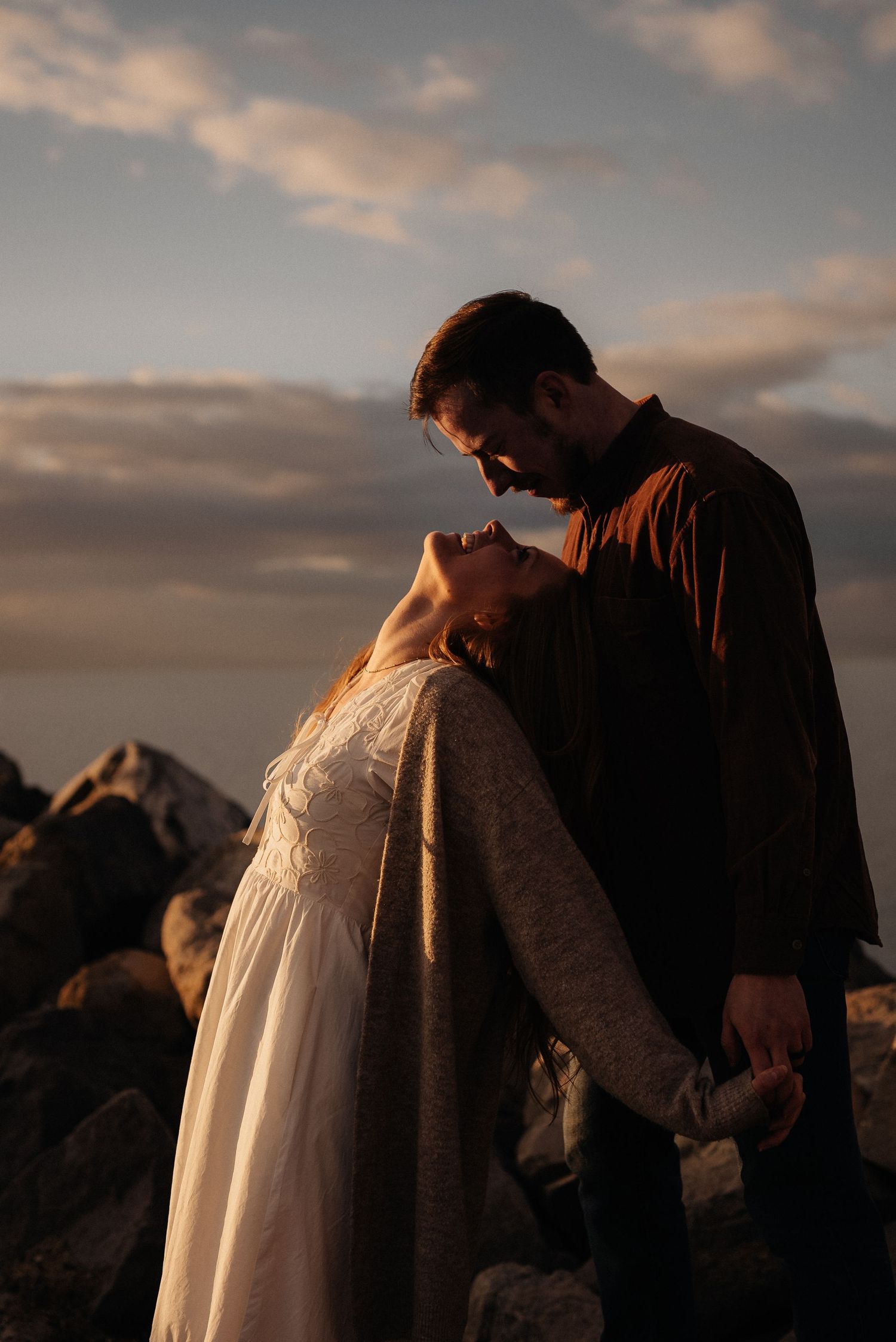 Silhouetted couple in wedding attire against dramatic sunset sky on rocky shore.