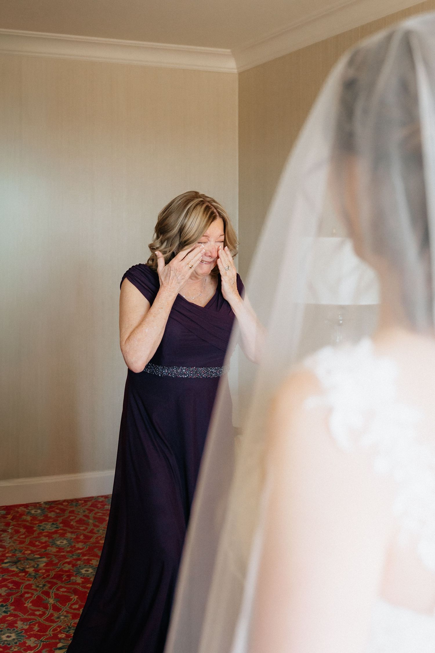 A tearful woman in a purple gown wipes her eye while seeing a bride in a white wedding dress and veil for the first time.