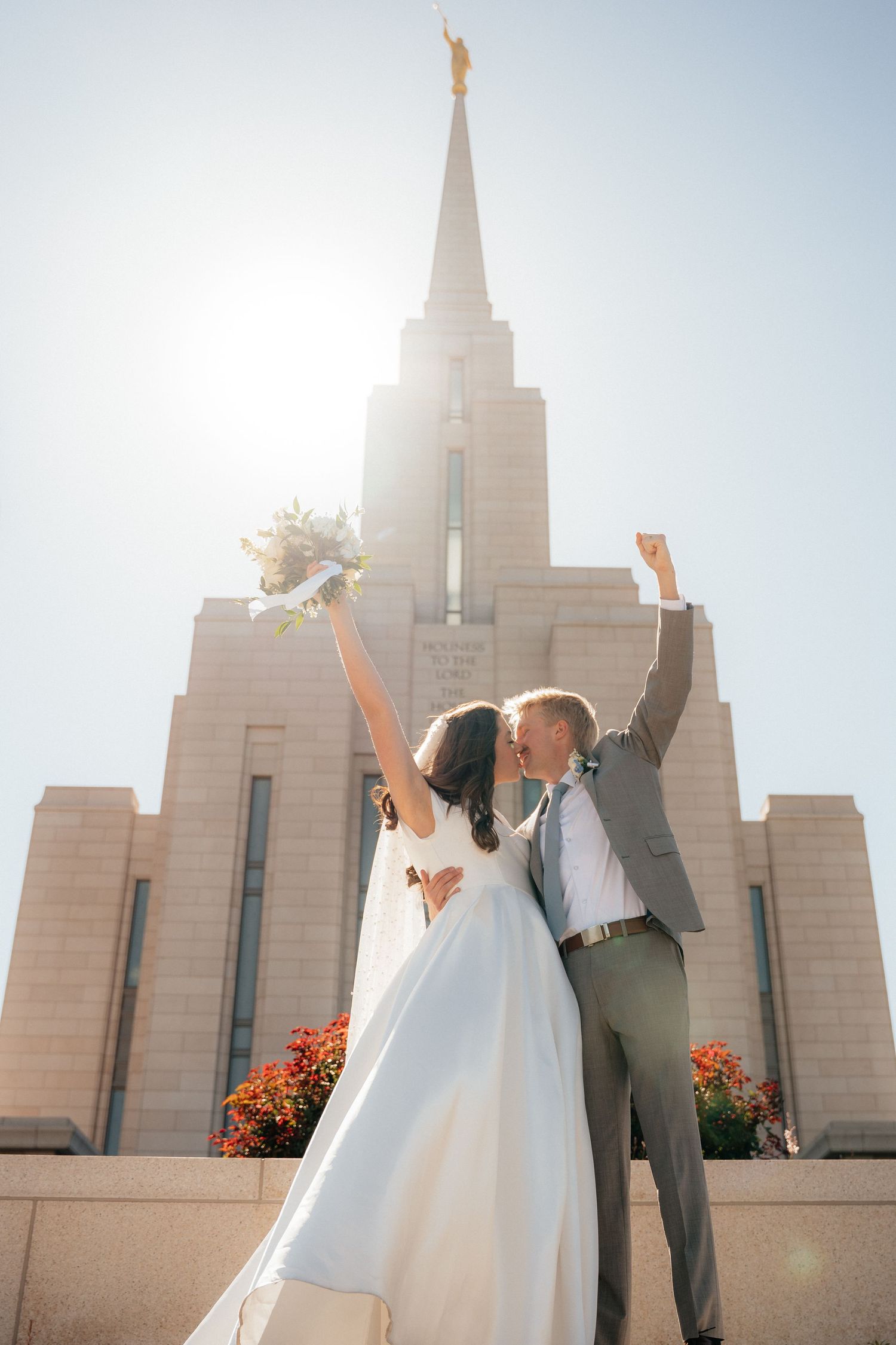 Bride and groom kissing and celebrating with raised arms in front of a sunlit LDS temple on their wedding day.