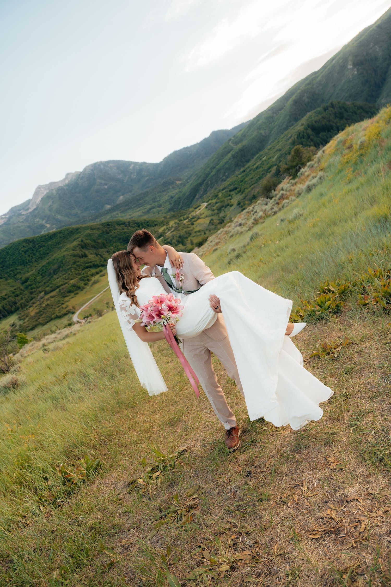 Groom dips bride for a kiss on a green mountain hillside, her white dress and pink bouquet flowing in the breeze.