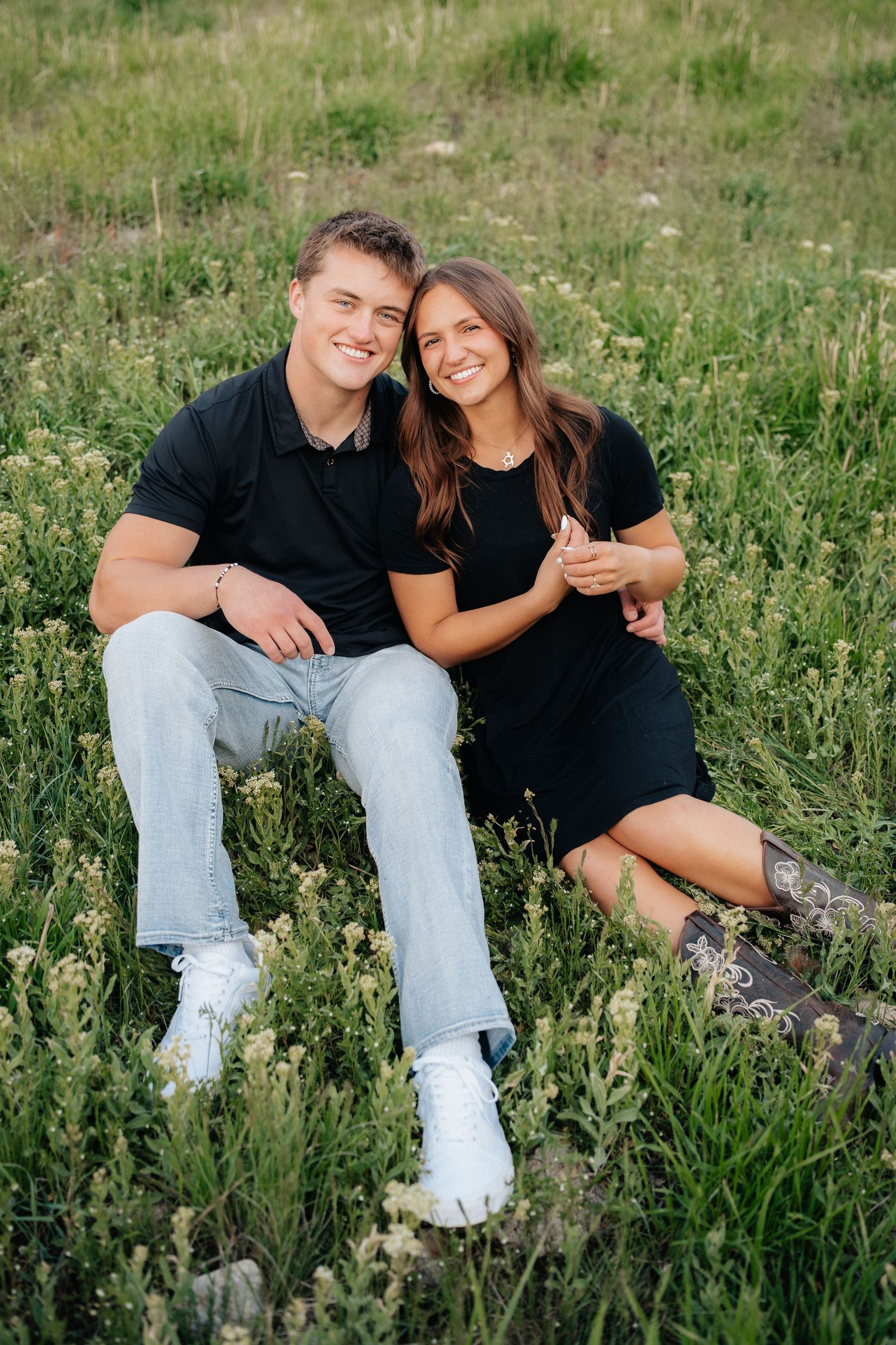 Happy young couple sitting together in a lush green meadow, both wearing black outfits, smiling warmly.