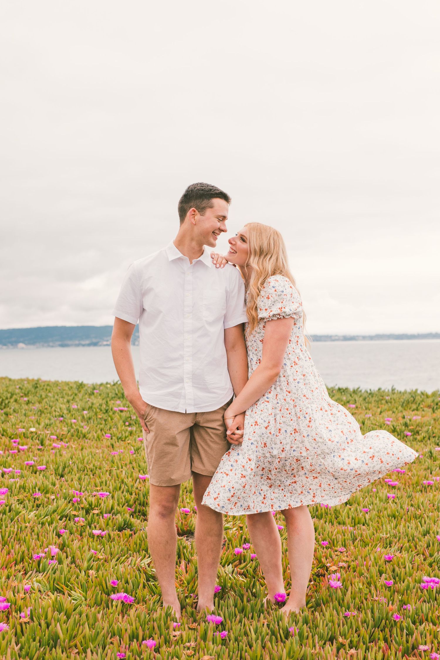 Couple in white dress and khaki shorts dancing in field of pink flowers by the ocean.