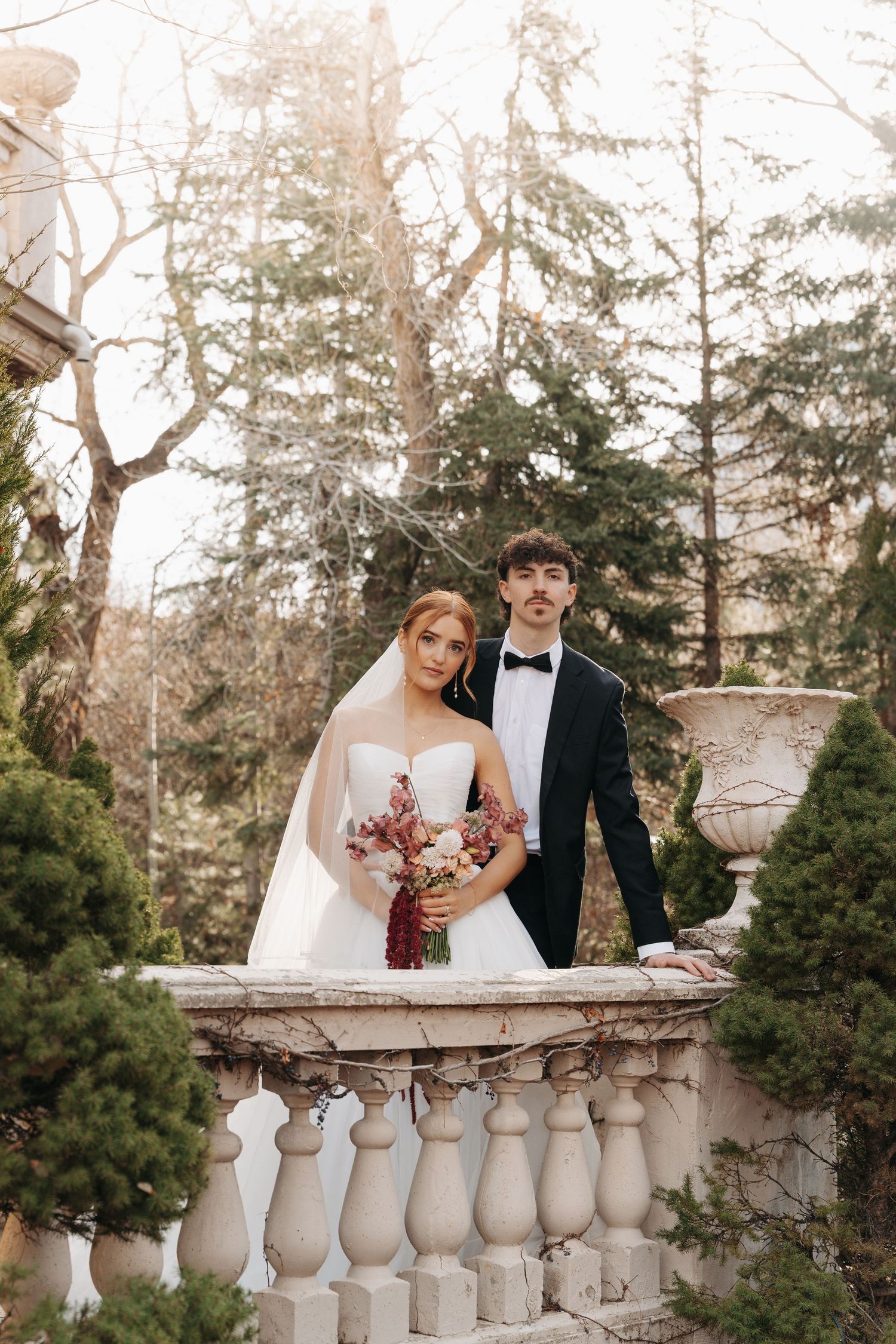 Wedding couple poses along ornate stone balustrade in garden setting during golden hour.