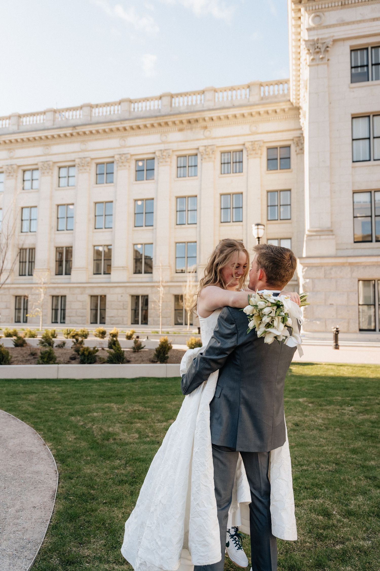 Groom lifting bride in white dress outside grand neoclassical building, holding white floral bouquet on sunny wedding day.
