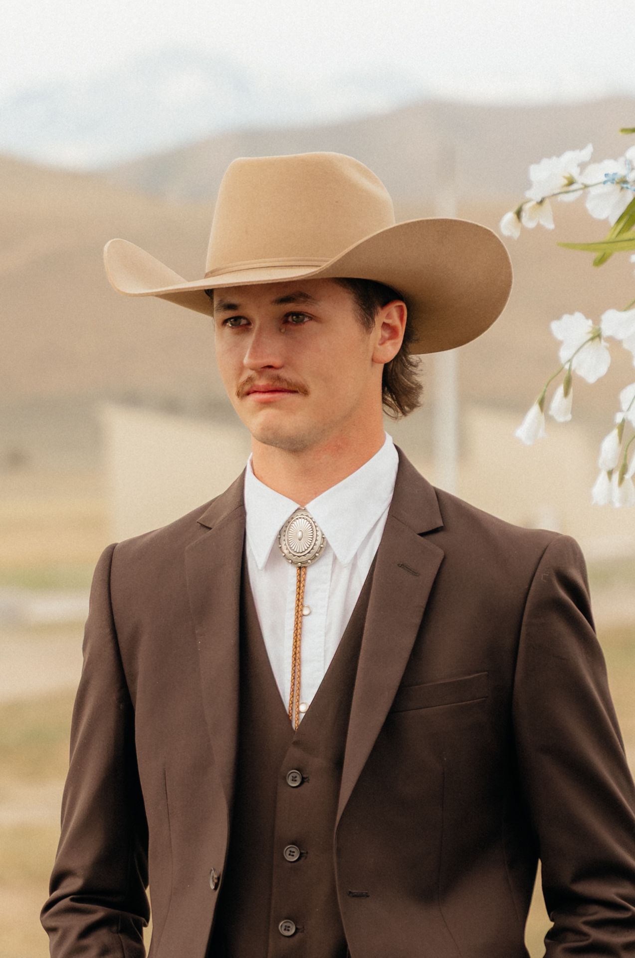 Young man in brown three-piece suit, bolo tie, and tan cowboy hat poses outdoors with mountain backdrop.