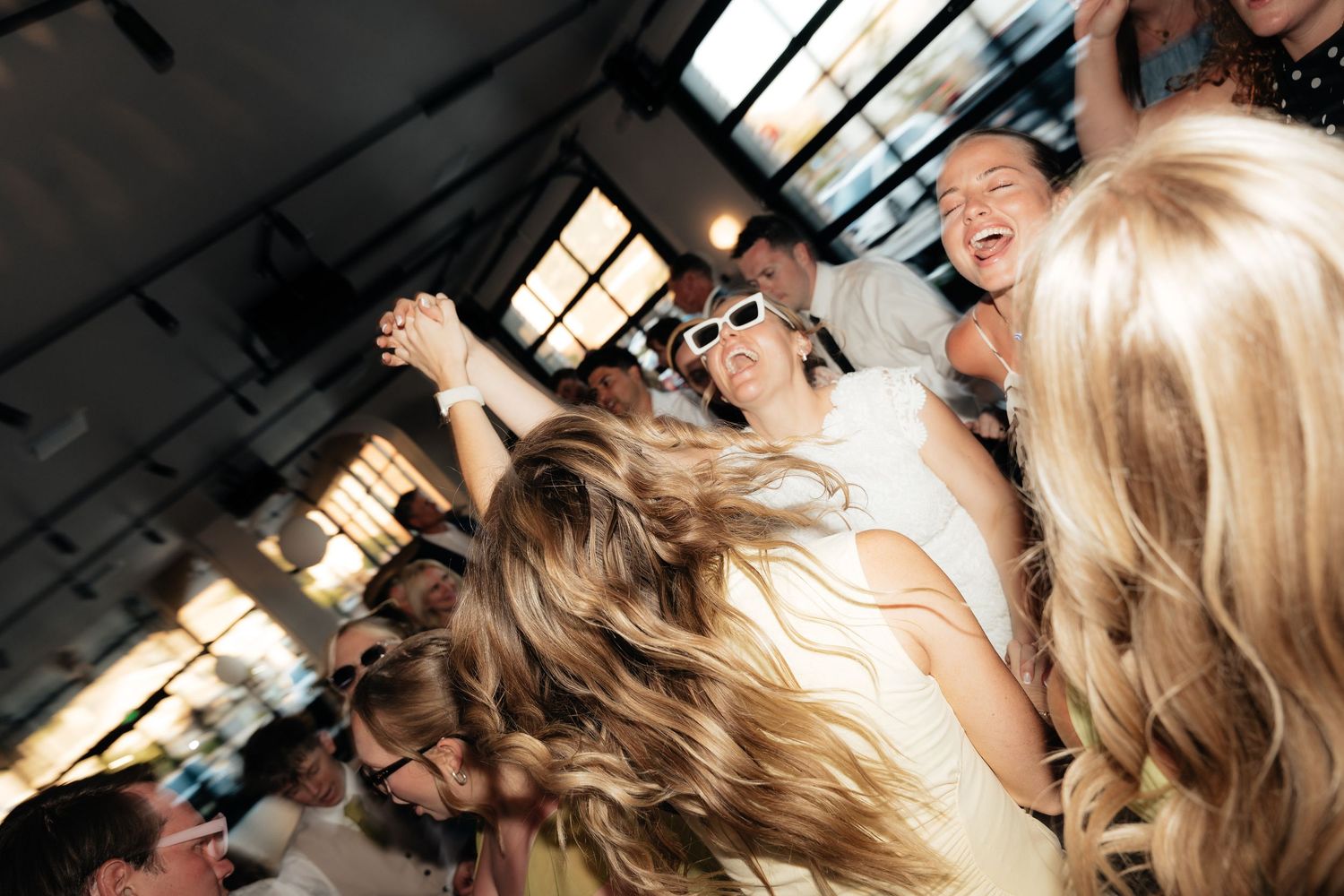 Joyful wedding reception crowd dancing and celebrating, bride in white dress with sunglasses laughing.