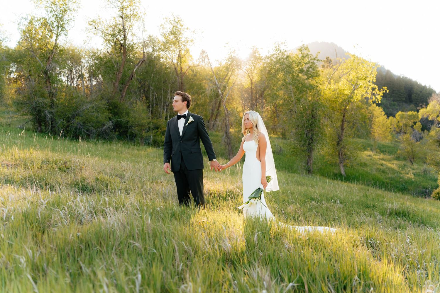 Bride and groom holding hands walking through a sunlit green meadow with trees in the background at golden hour.