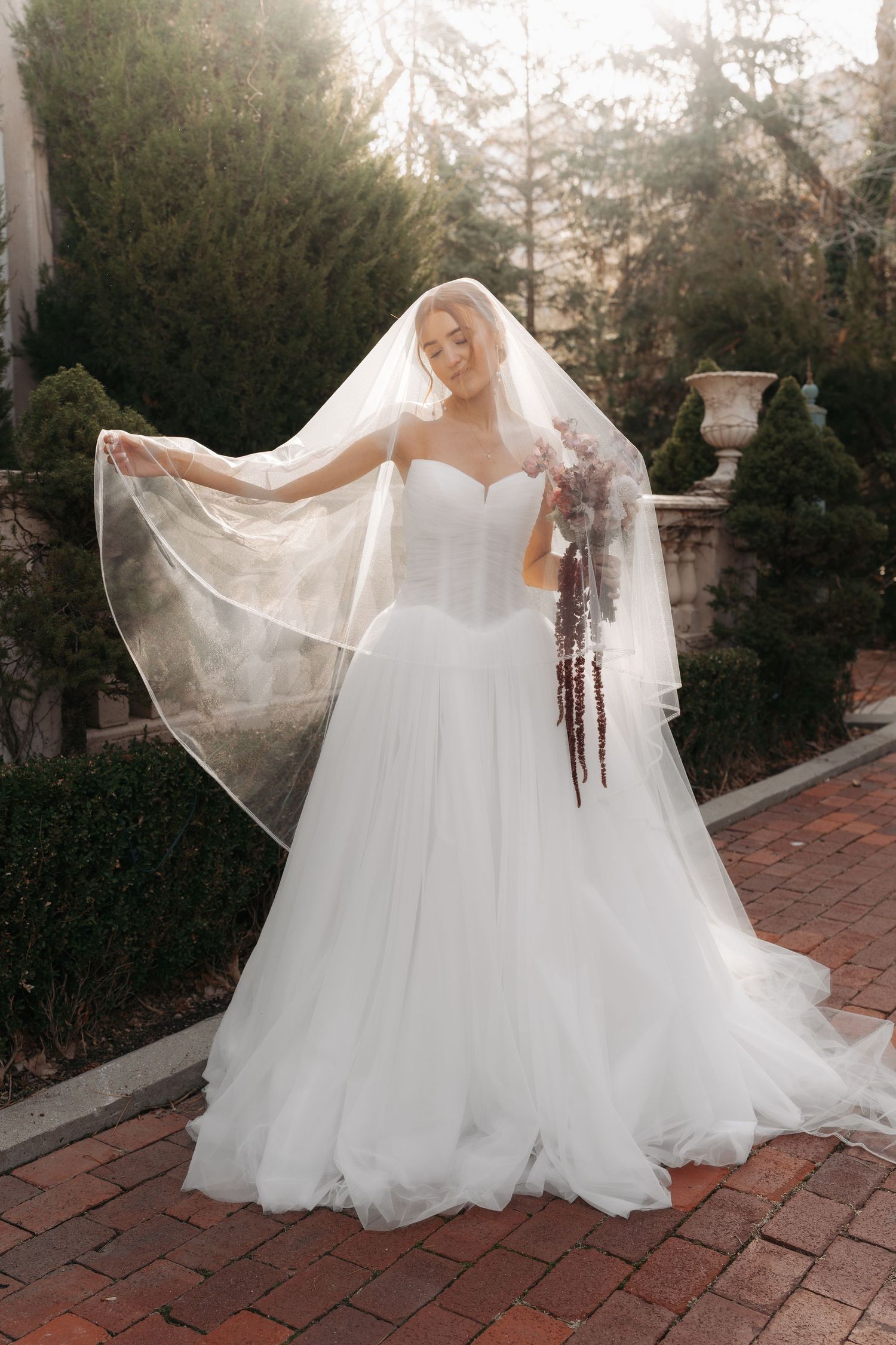 A bride in a flowing white tulle gown and veil poses on a brick pathway surrounded by garden greenery at sunset.