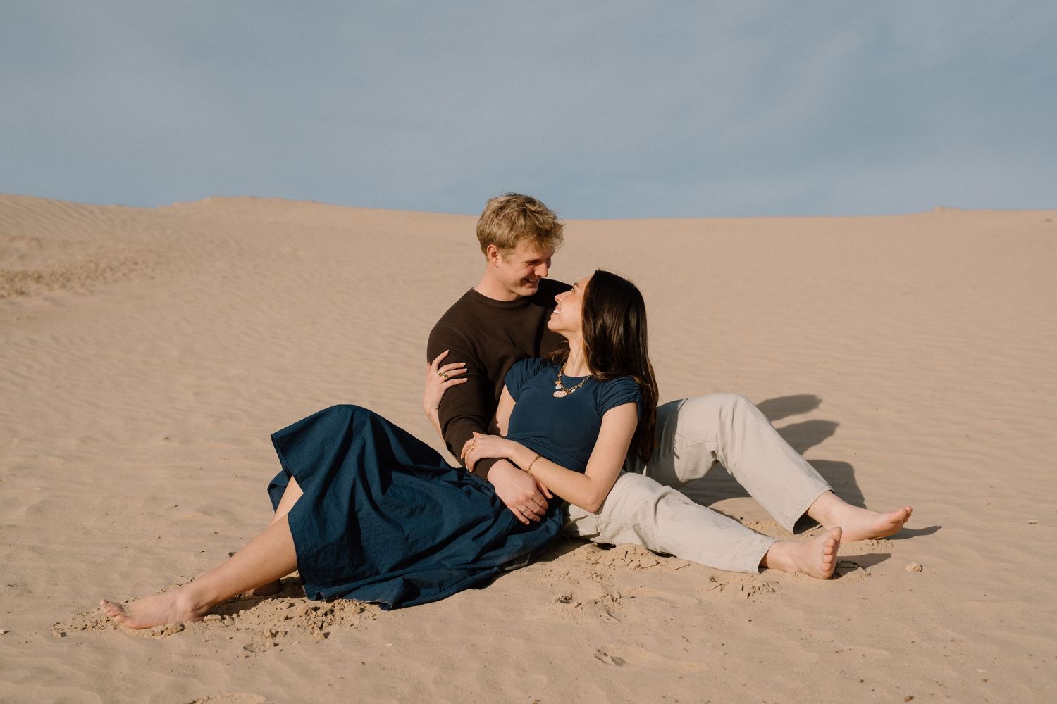 A romantic couple in blue and white outfits lying together on sandy beach dunes during sunset.