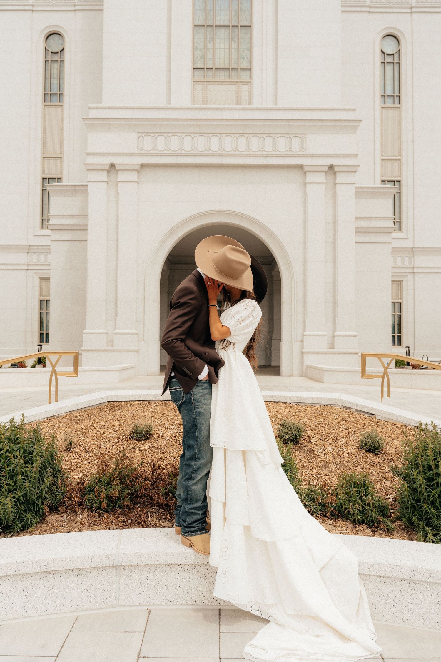 Bride and groom sharing a kiss outside a white LDS temple, groom wearing a tan cowboy hat and jeans.