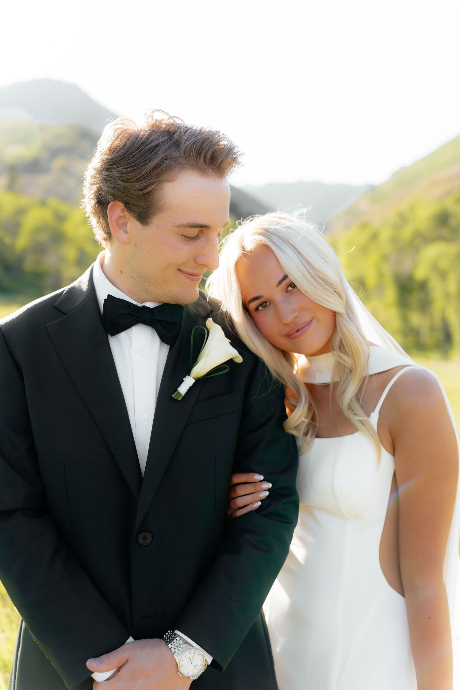 Bride and groom in black tuxedo and white dress pose together outdoors with green hills in background.
