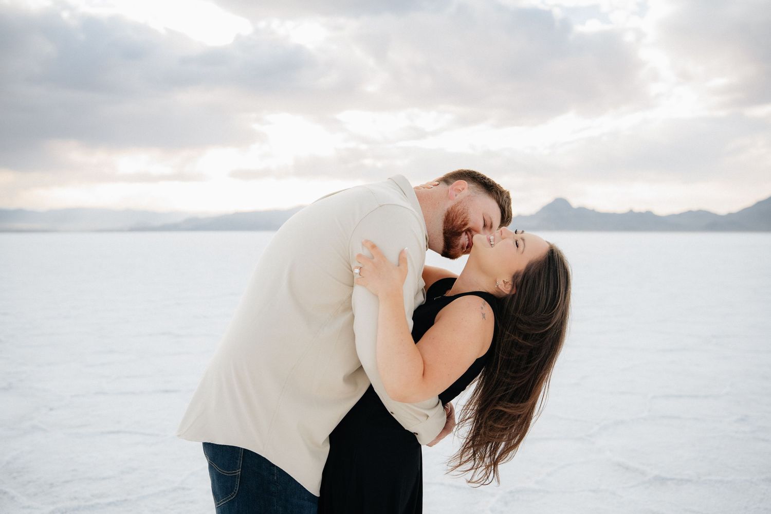 Couple sharing a romantic dip kiss on the Bonneville Salt Flats with mountains and cloudy sky backdrop.
