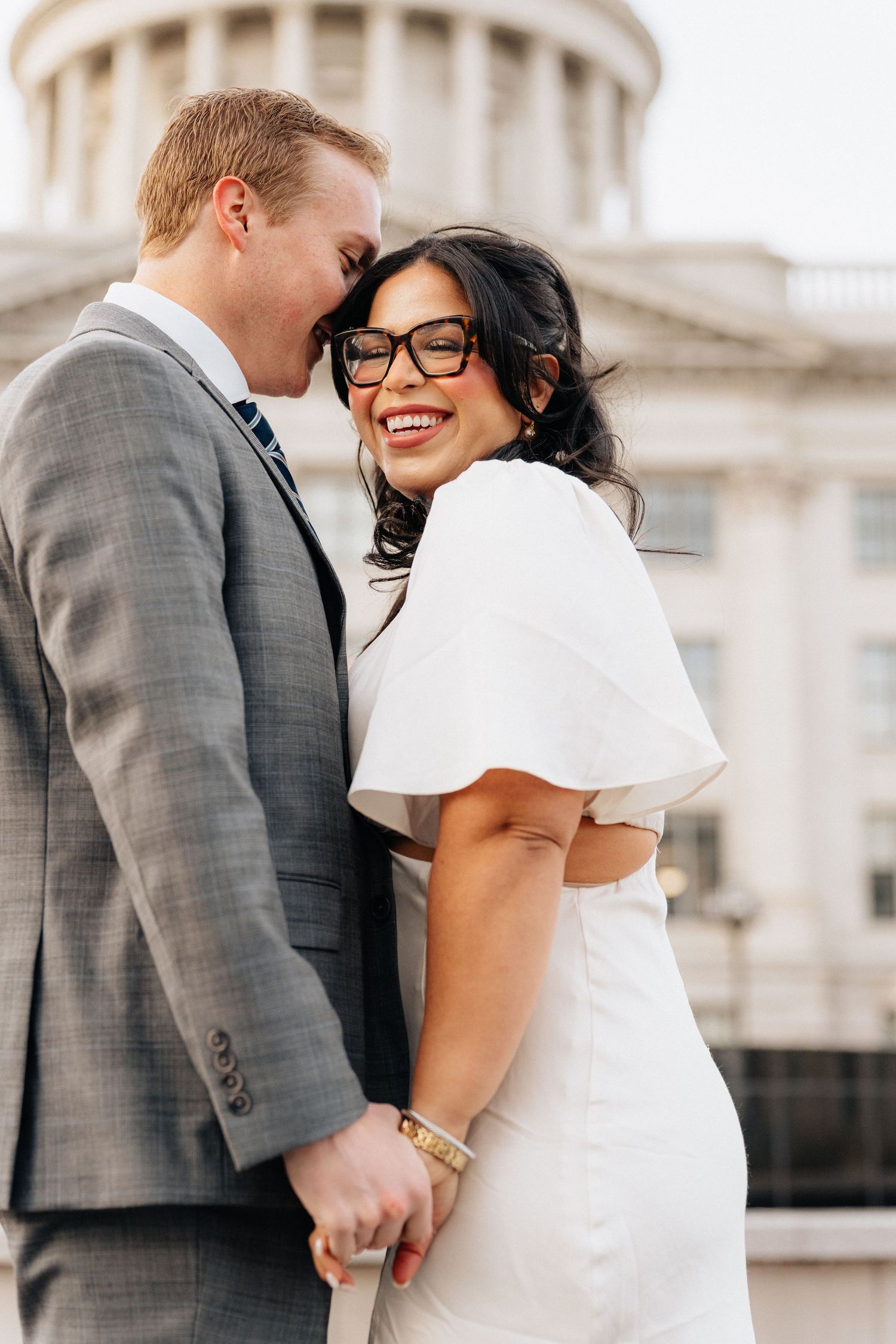 Happy couple in wedding attire holding hands, bride in white dress and glasses smiling, groom in gray suit, capitol building backdrop.
